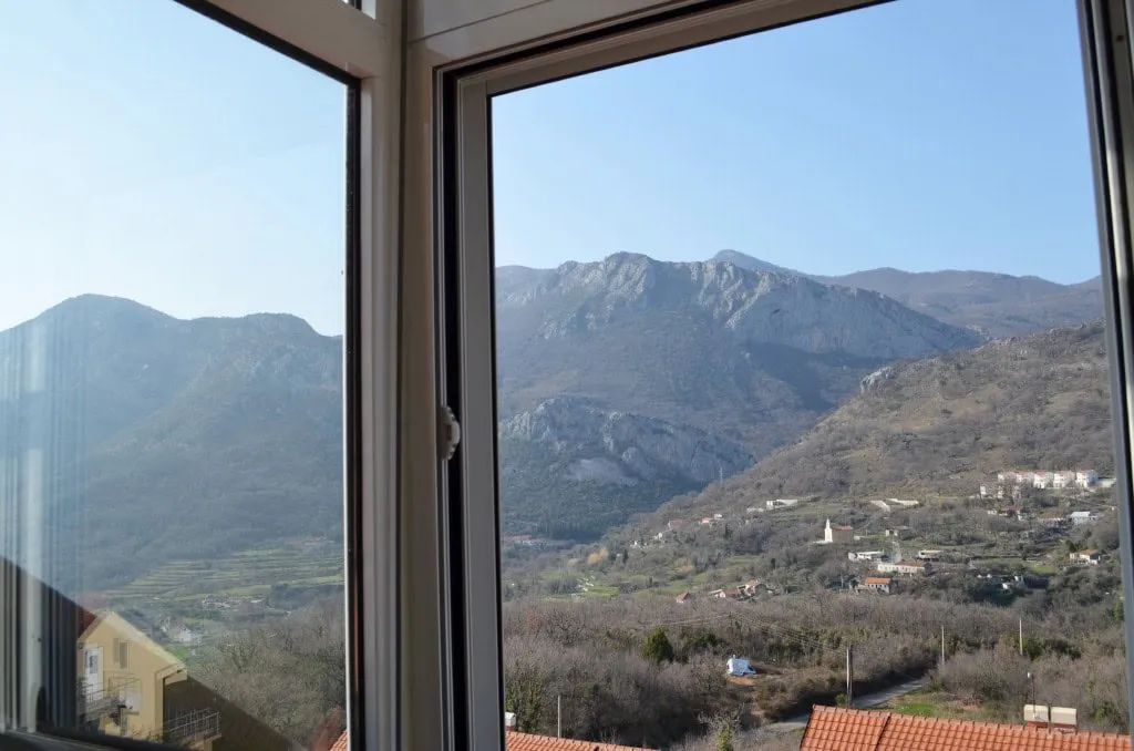 View through a white window frame showing mountains, a village, and tiled rooftops below.
