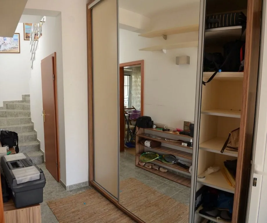 Hallway with sliding wardrobe, staircase, and door. Floor mat visible; shelves hold bags, sports gear, and miscellaneous items.