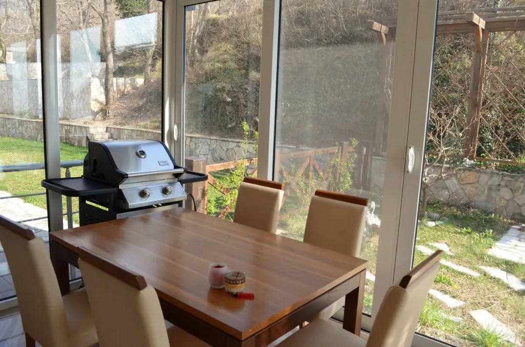 Glassed-in terrace with a wooden dining table, chairs, and a gas grill overlooking a garden.