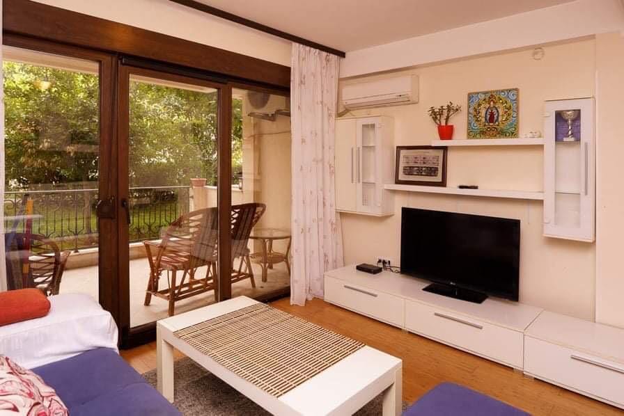 Living room with access to the terrace in an apartment in Budva, Montenegro: light furniture, TV area, and a view of the green area.