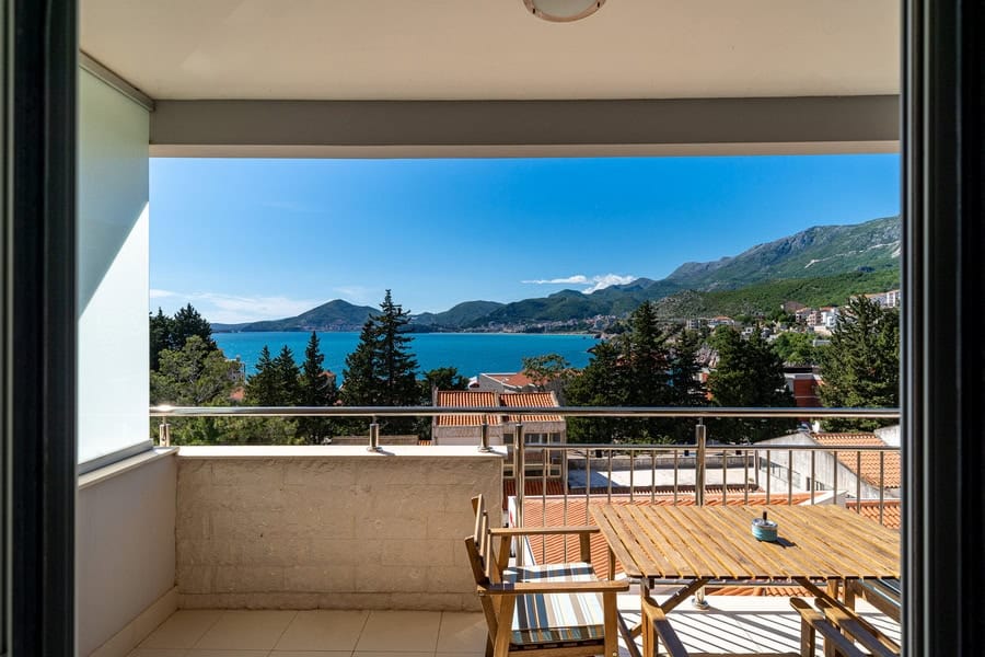 Balcony with wooden furniture and metal railing overlooking the sea and mountains.