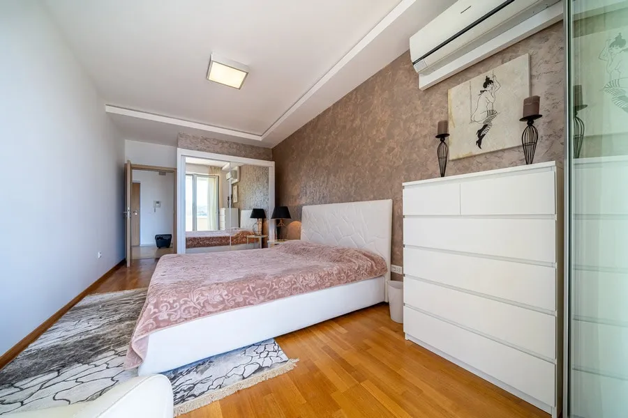 Modern bedroom featuring a white bed with pink cover, white dresser, wooden floor, and patterned rug.