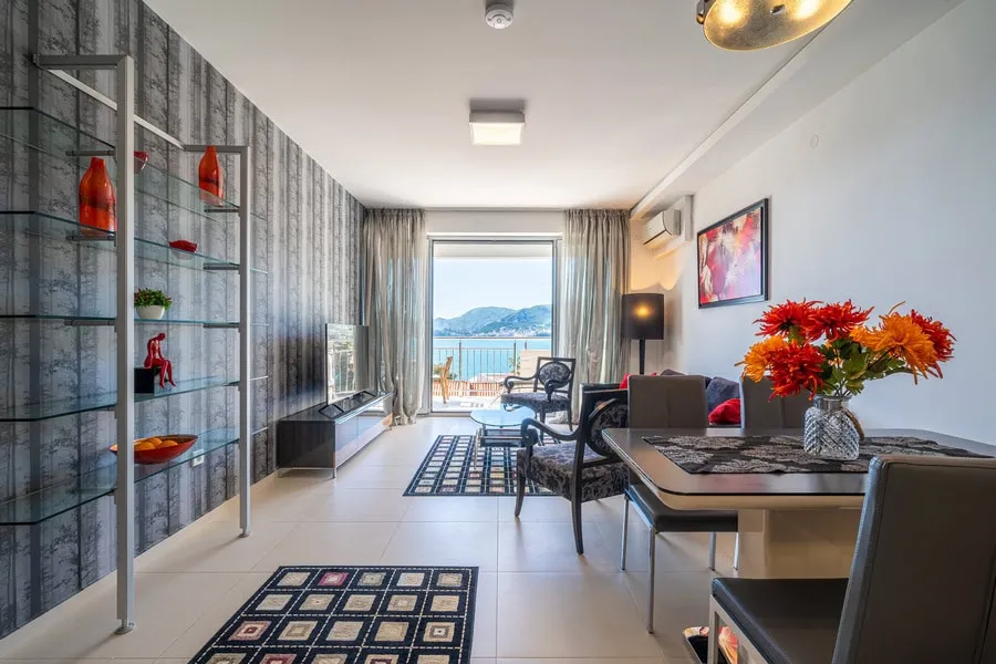 Living room featuring a sofa, coffee table, TV unit, dining area, and glass shelves overlooking the sea through balcony doors.