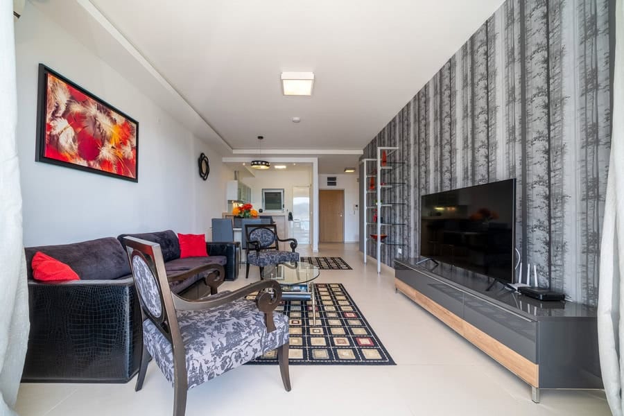 Living room with sofa, armchair, TV on console, and patterned rug. Features tree-motif wallpaper and wall art.