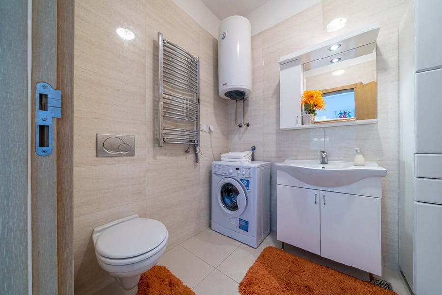Bathroom featuring a toilet, sink vanity, washing machine, wall-mounted water heater, and heated towel rail on beige tiled walls.