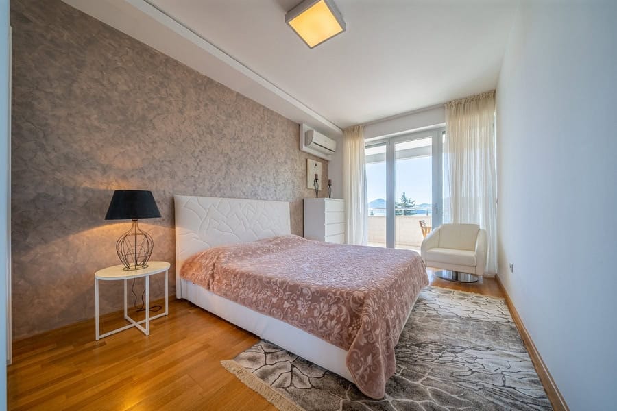 Bedroom featuring a double bed with white headboard, wooden flooring, side table, and balcony door overlooking the sea.