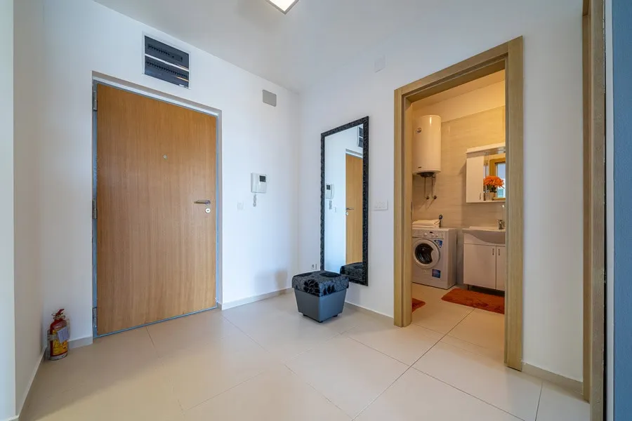 Entryway with a wooden door, mirror, and ottoman; an adjacent room shows a washing machine and sink.