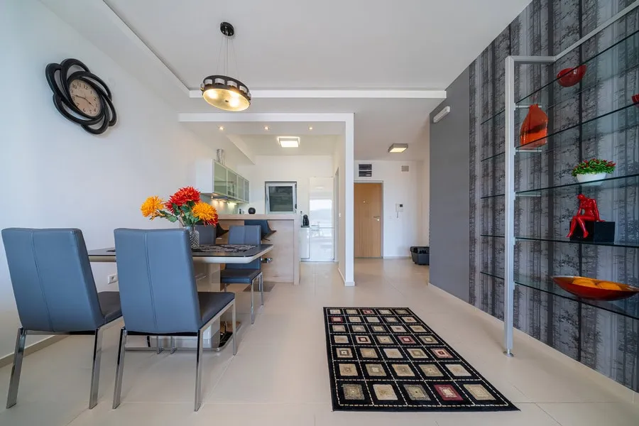 Modern open-plan living and dining area with blue chairs, a glass shelving unit, patterned rug, and kitchen visible in the background.