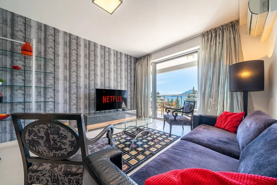 Living room with blue sofa, Netflix TV, glass table, and balcony door overlooking the sea and mountains.