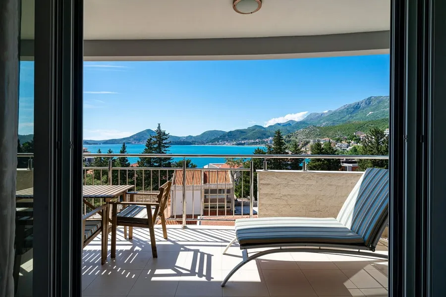 Balcony with wooden furniture and lounge chair overlooking the sea and mountains.