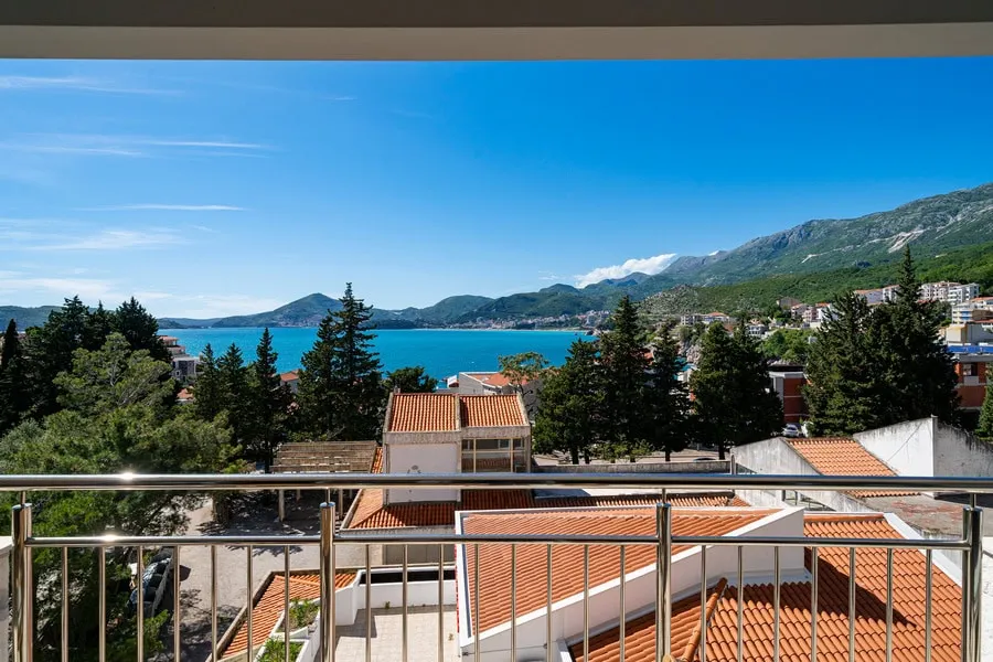 View from a balcony with metal railing overlooking sea, mountains, and rooftops.