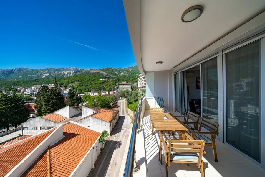 Balcony with wooden dining set overlooking tiled rooftops and green mountains under a clear blue sky.