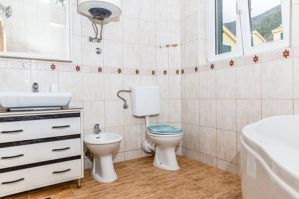 Bright bathroom in an apartment in Đenovići, Montenegro: vanity with sink, bidet, toilet, and mountain view from the window.