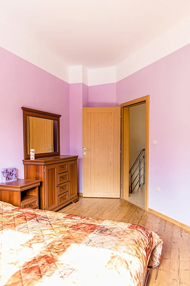 Bedroom with pink walls, wooden furniture including a dresser with mirror, and a bed with patterned quilt on wood floor.