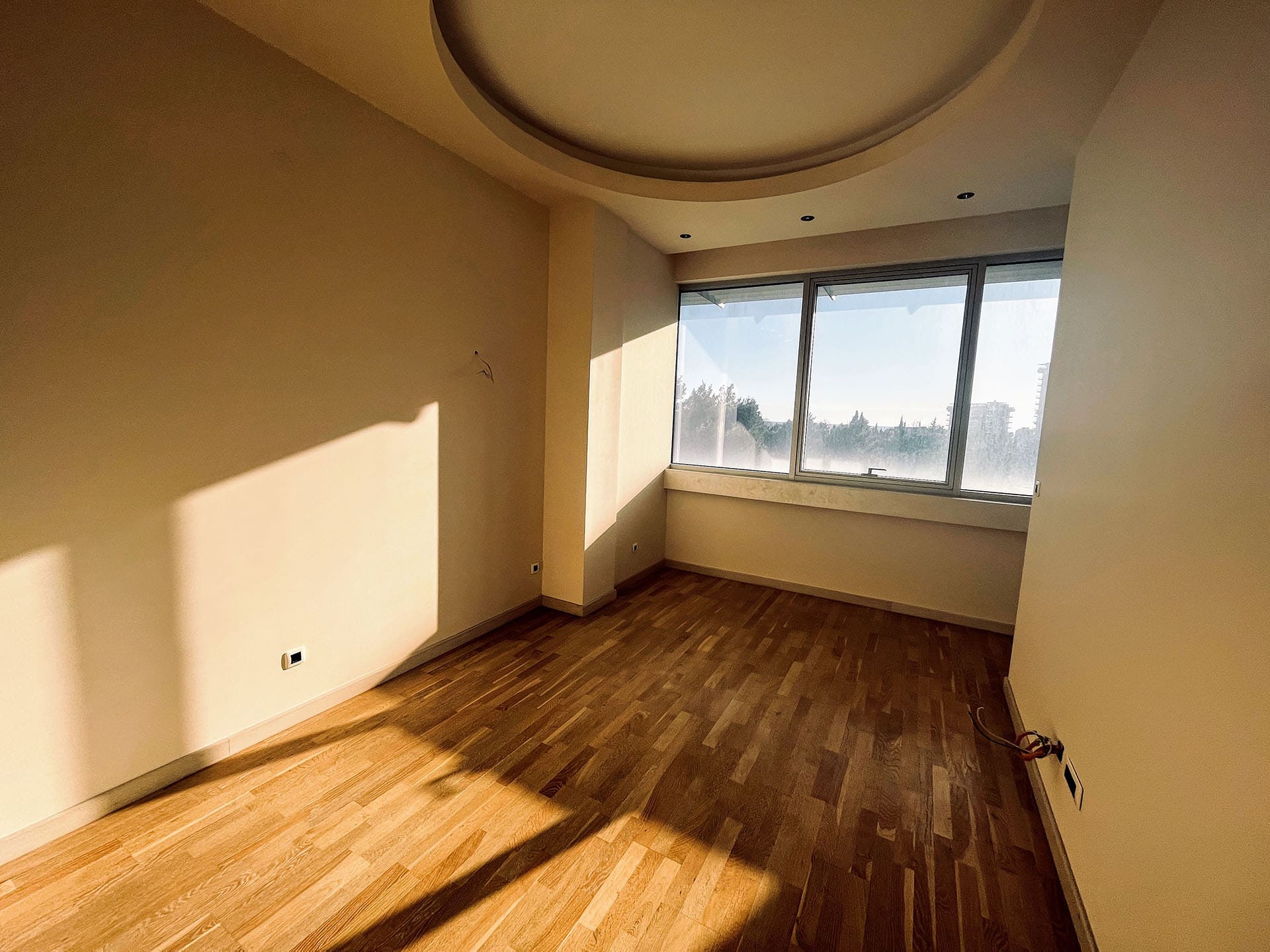 Bright living room in an apartment in Budva, Montenegro: parquet floor, panoramic window, and a multi-level ceiling with lighting.