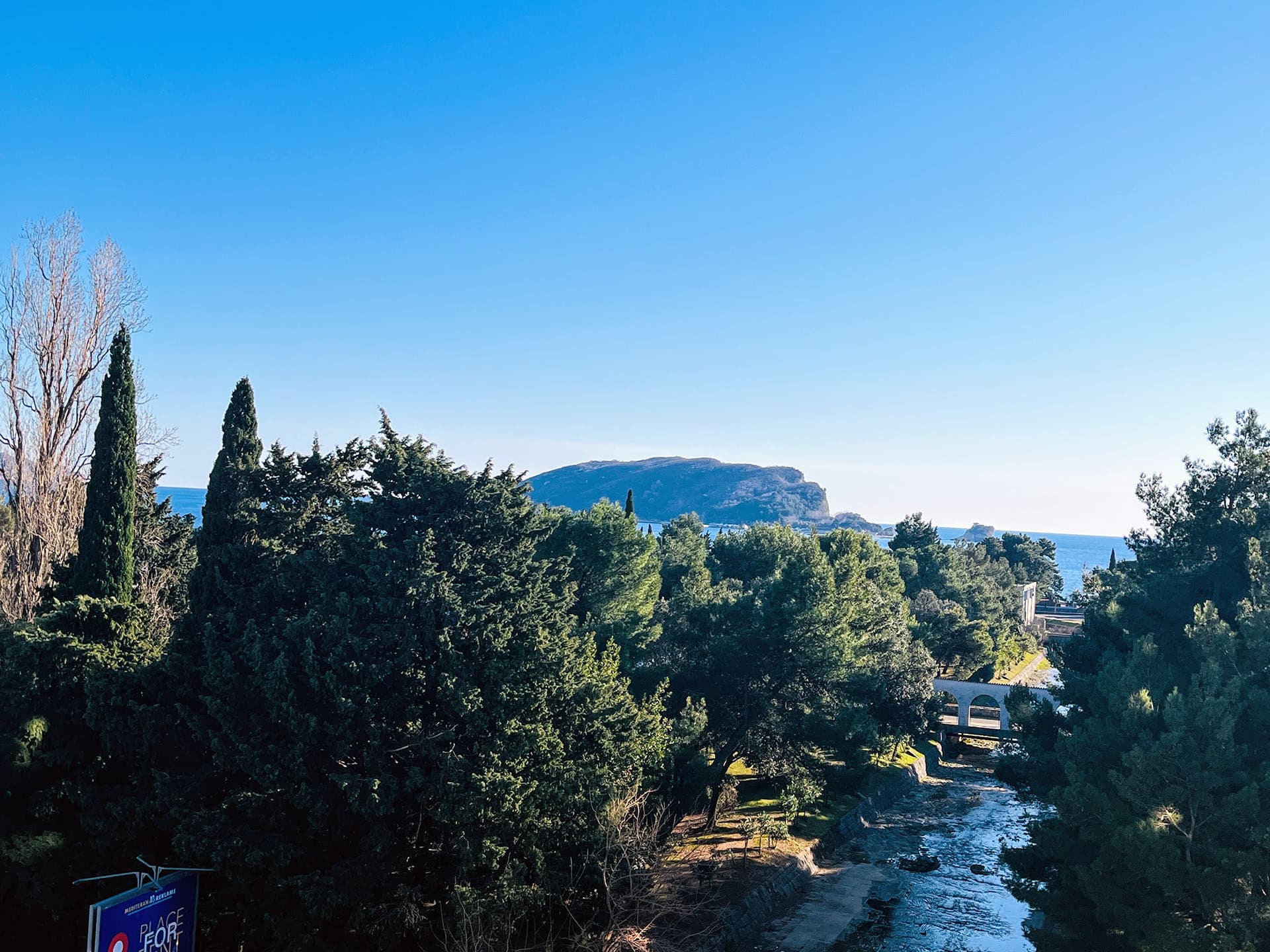 Panoramic view of the sea, mountains, and river in Budva, Montenegro, with green trees and a pedestrian bridge in the foreground.