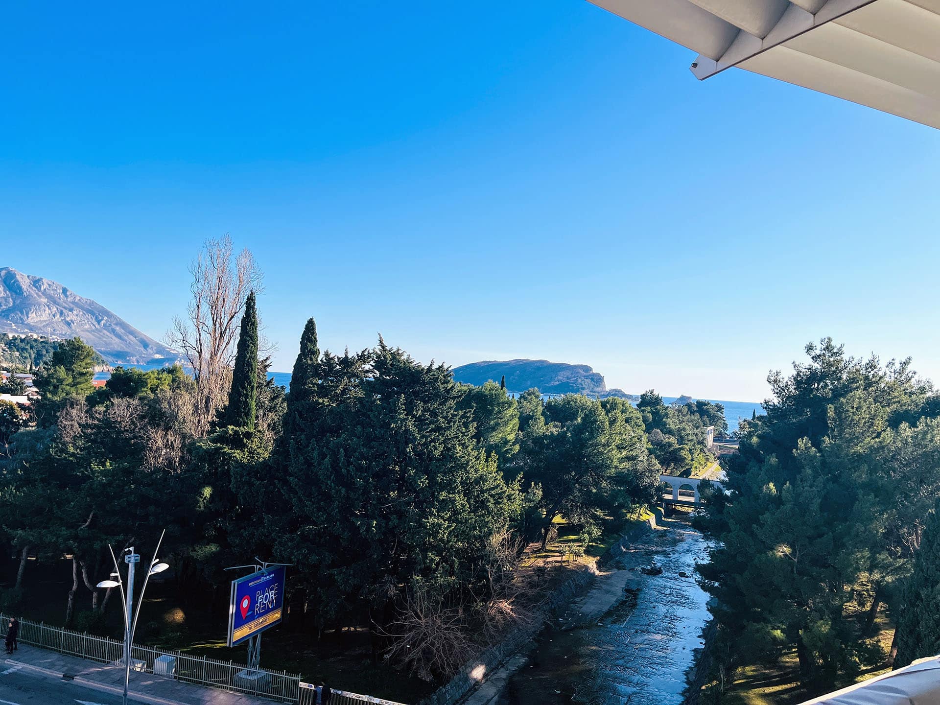 View from the apartment balcony in Budva, Montenegro: a panorama of mountains, a green park, a river, and the seaside.