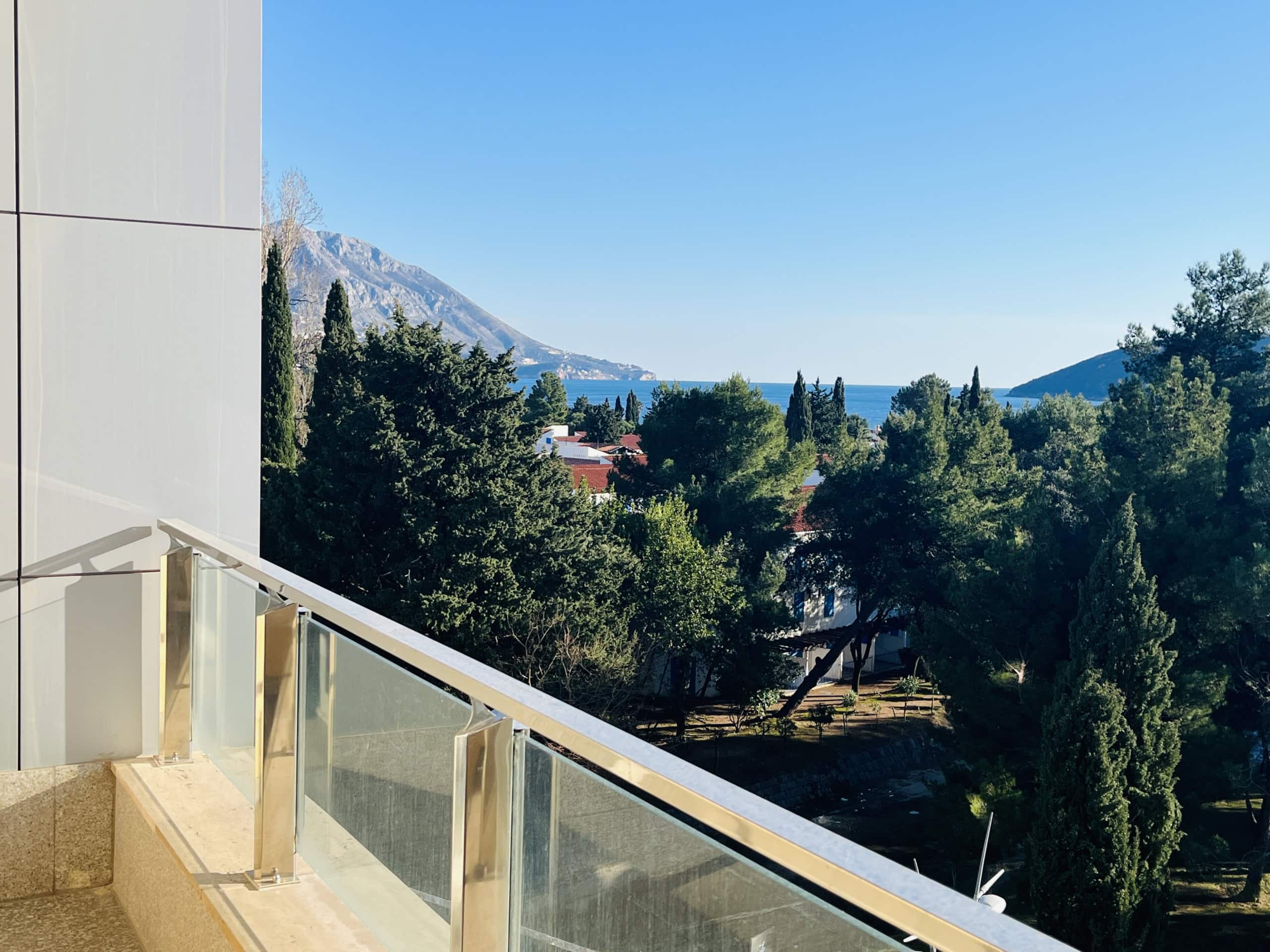 View from the apartment balcony in Budva, Montenegro: a panorama of the sea, mountains, and a green park with conifer trees.