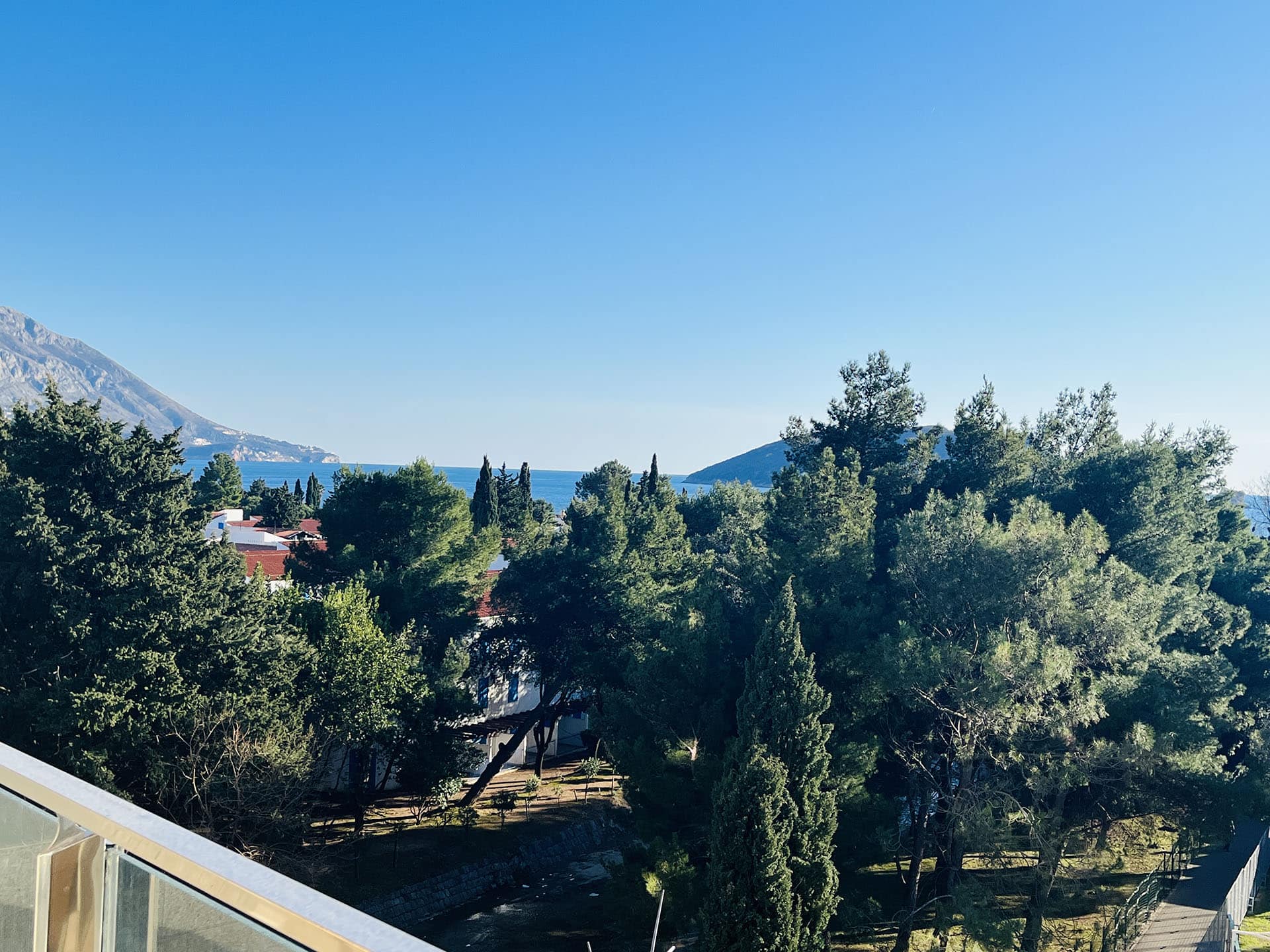 View from the apartment balcony in Budva, Montenegro: a panorama of the Adriatic Sea, a green park, and mountains on a sunny day.