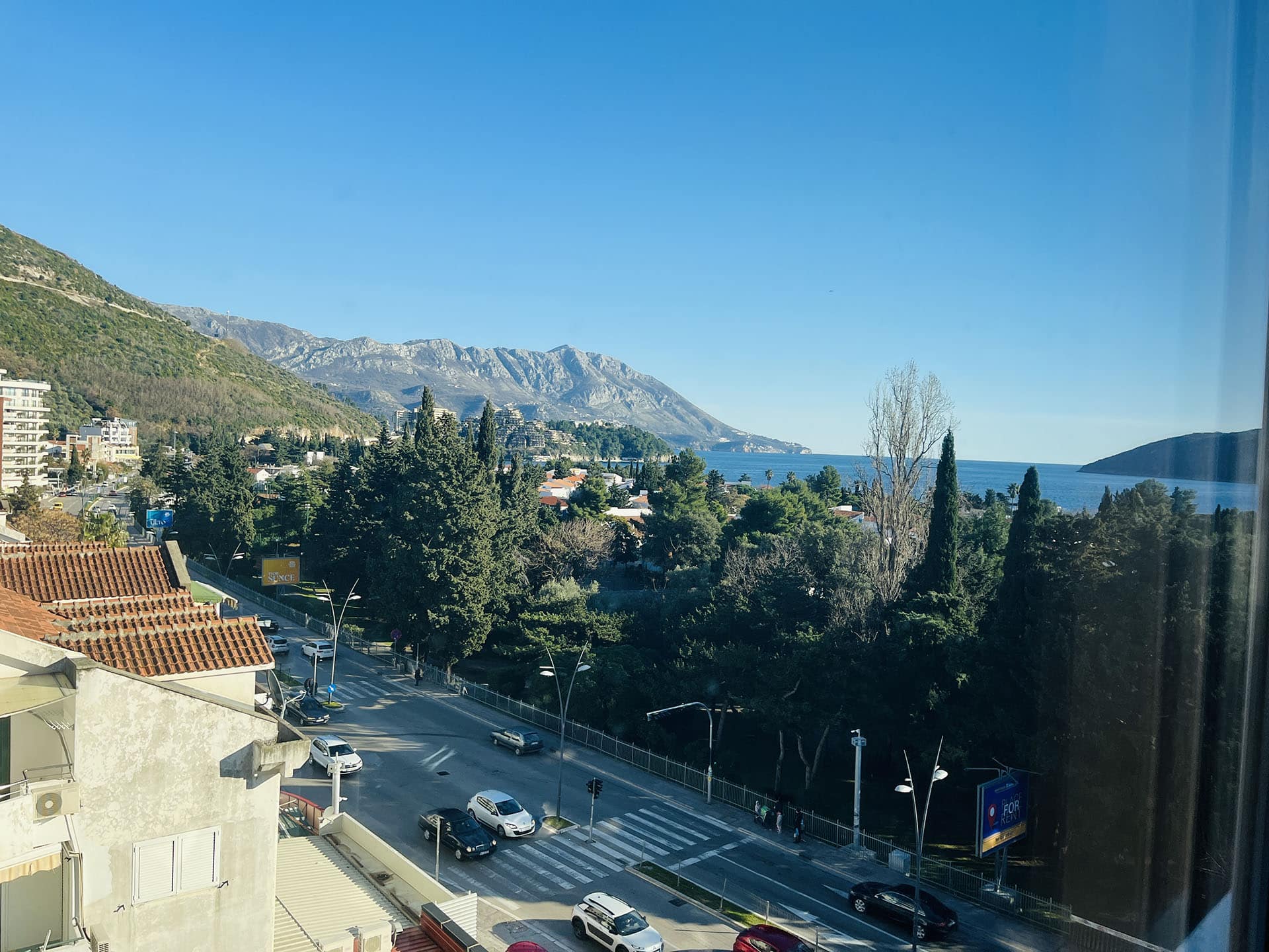 View from the apartment balcony in Budva, Montenegro: a panorama of the sea, mountains, greenery, and a city street on a sunny day.