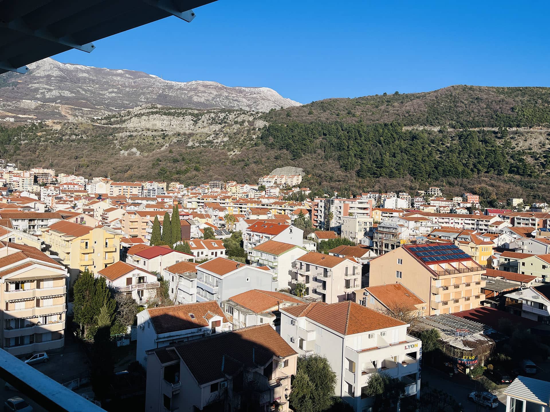 Panoramic view from the apartment balcony in Budva, Montenegro: city buildings with tiled roofs and a mountain landscape.