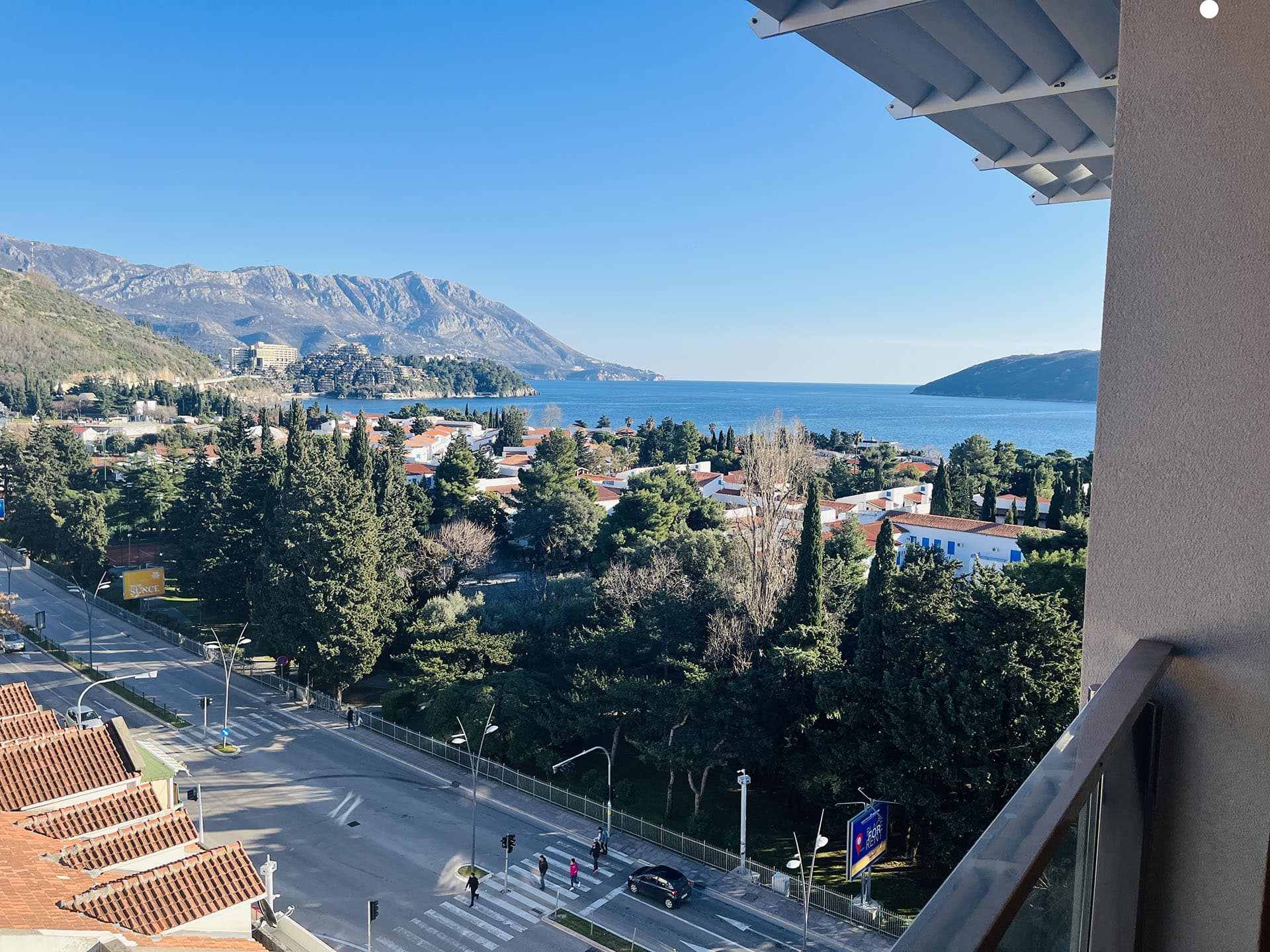 Panoramic view from the apartment balcony in Budva, Montenegro: sea, mountains, a green park, and city infrastructure.