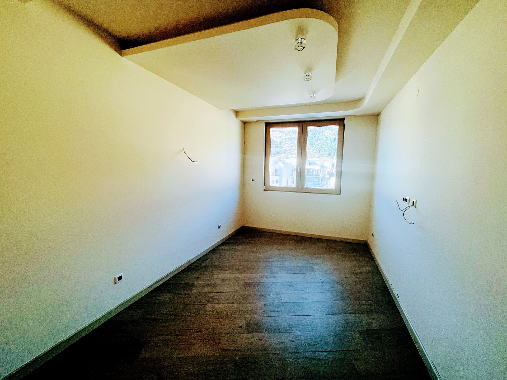 Bright living room in a new apartment in Budva, Montenegro: designer ceiling, dark laminate flooring, and a view from the window.