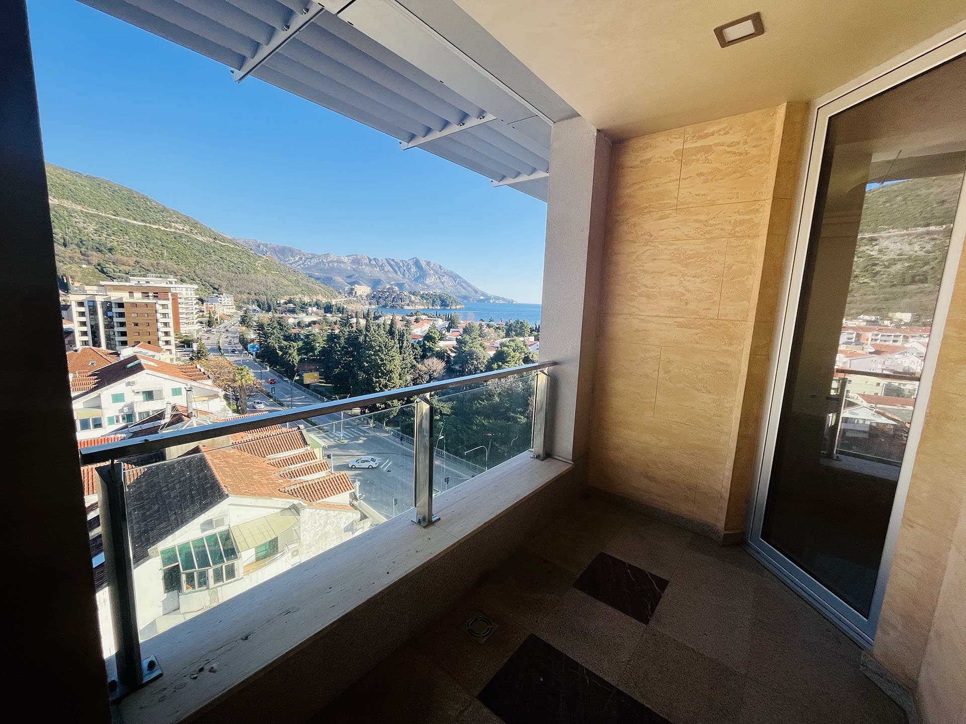 Apartment balcony in Budva, Montenegro, with panoramic views of the sea, mountains, and city, glass railing, and stone finishes.