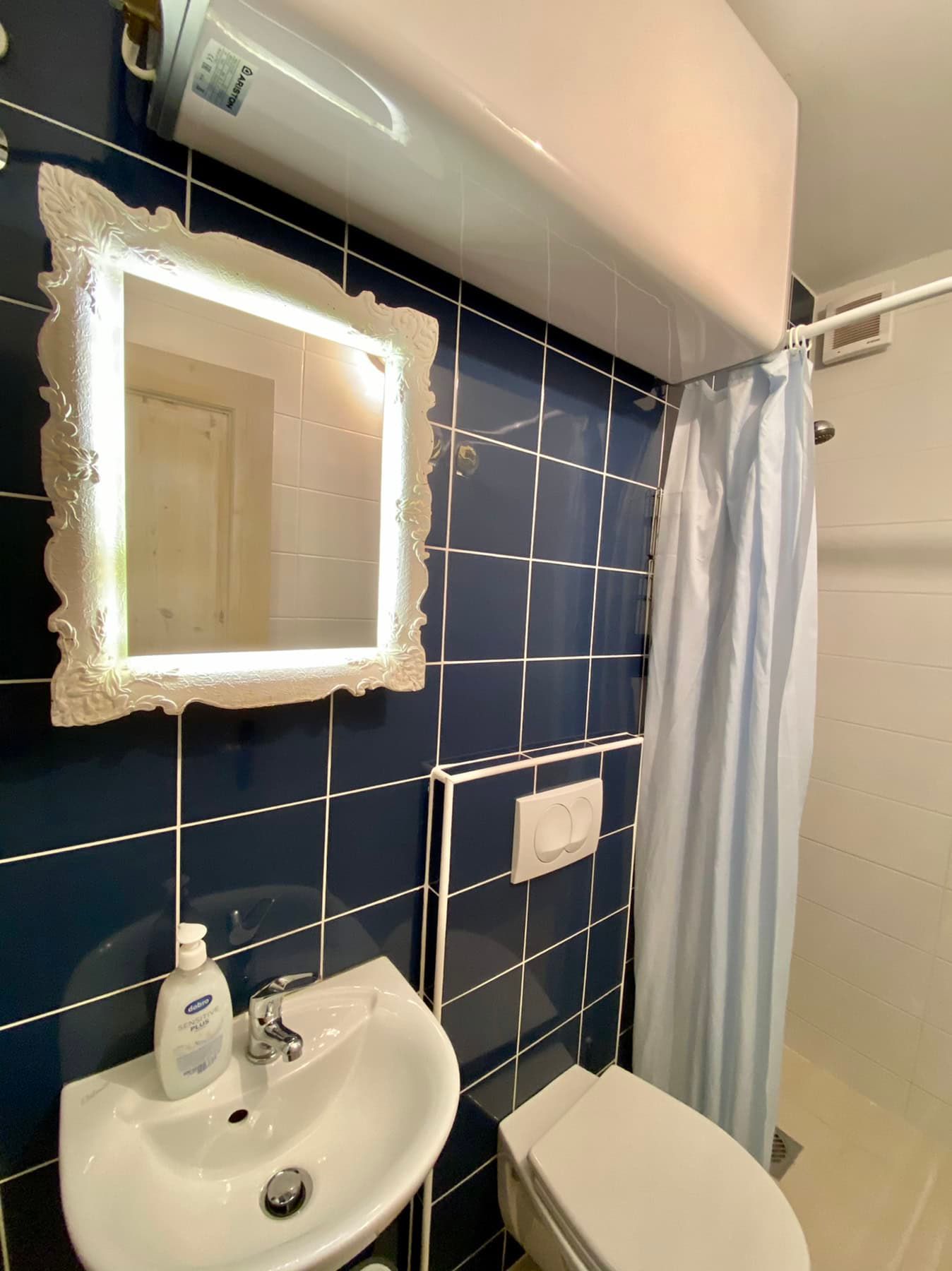 Bathroom in a house in Shushan, Montenegro: blue tiles, illuminated mirror, sink, and shower with curtain.