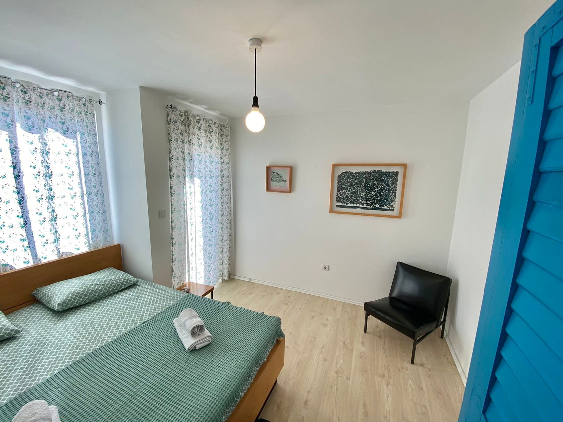 Bright bedroom in a house in Shushan, Montenegro: a double bed, black armchair, light floor, and a view of the terrace.