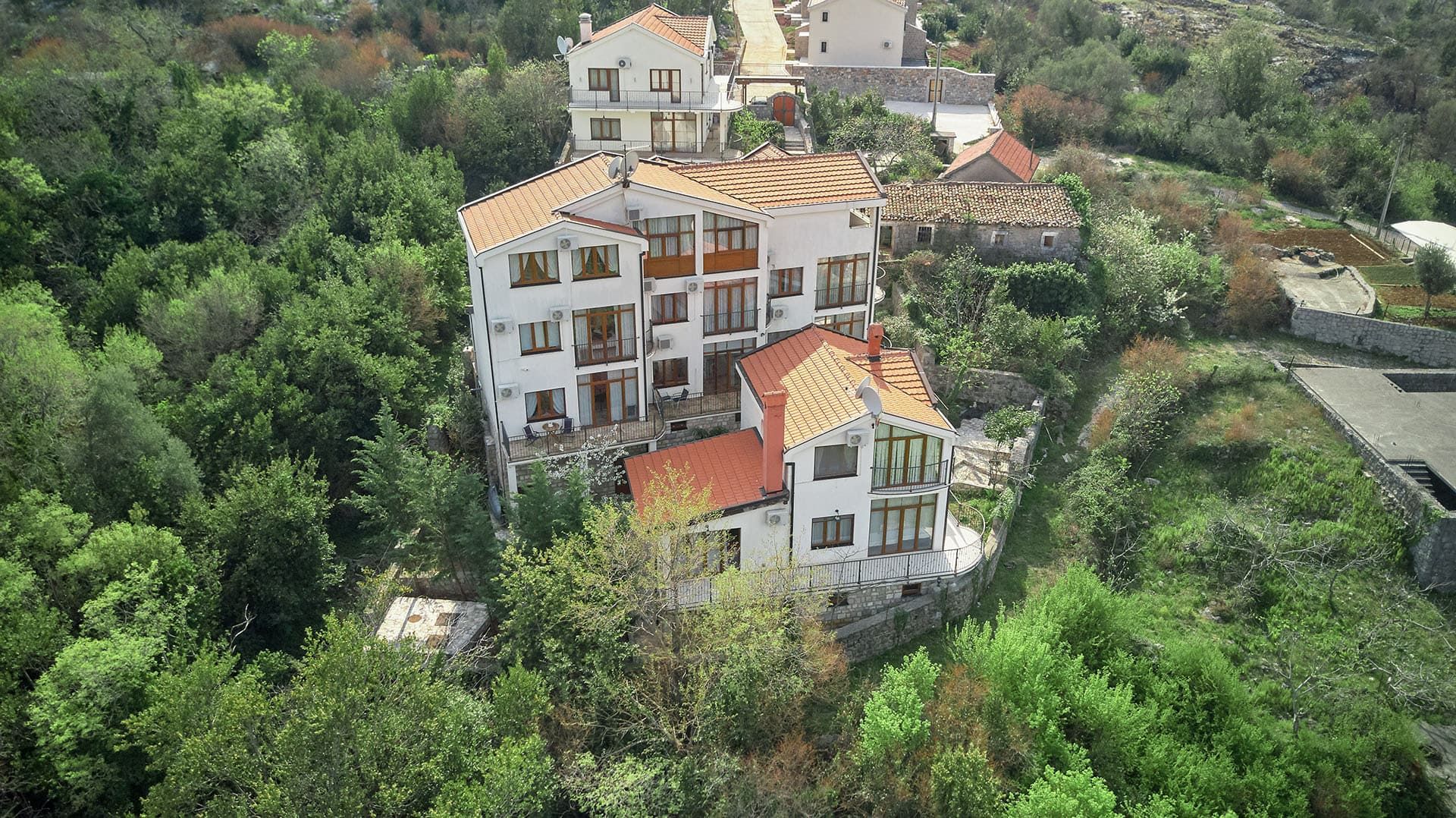 Aerial view of a white multi-story house with a red tiled roof, surrounded by dense green vegetation and trees on a hillside.