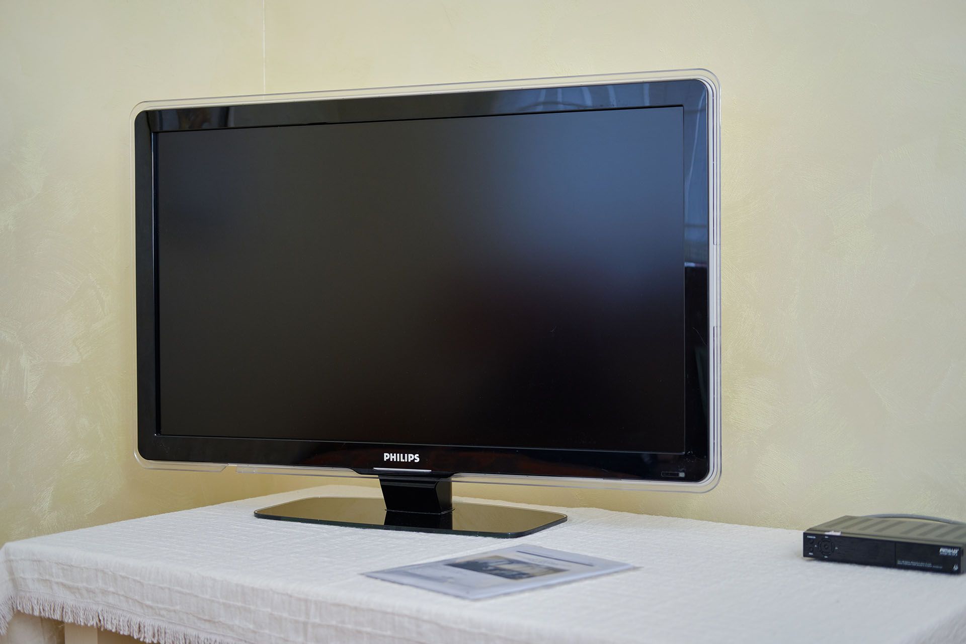 A flat-screen Philips TV sits on a table covered with a white cloth next to a black receiver against a yellow wall.