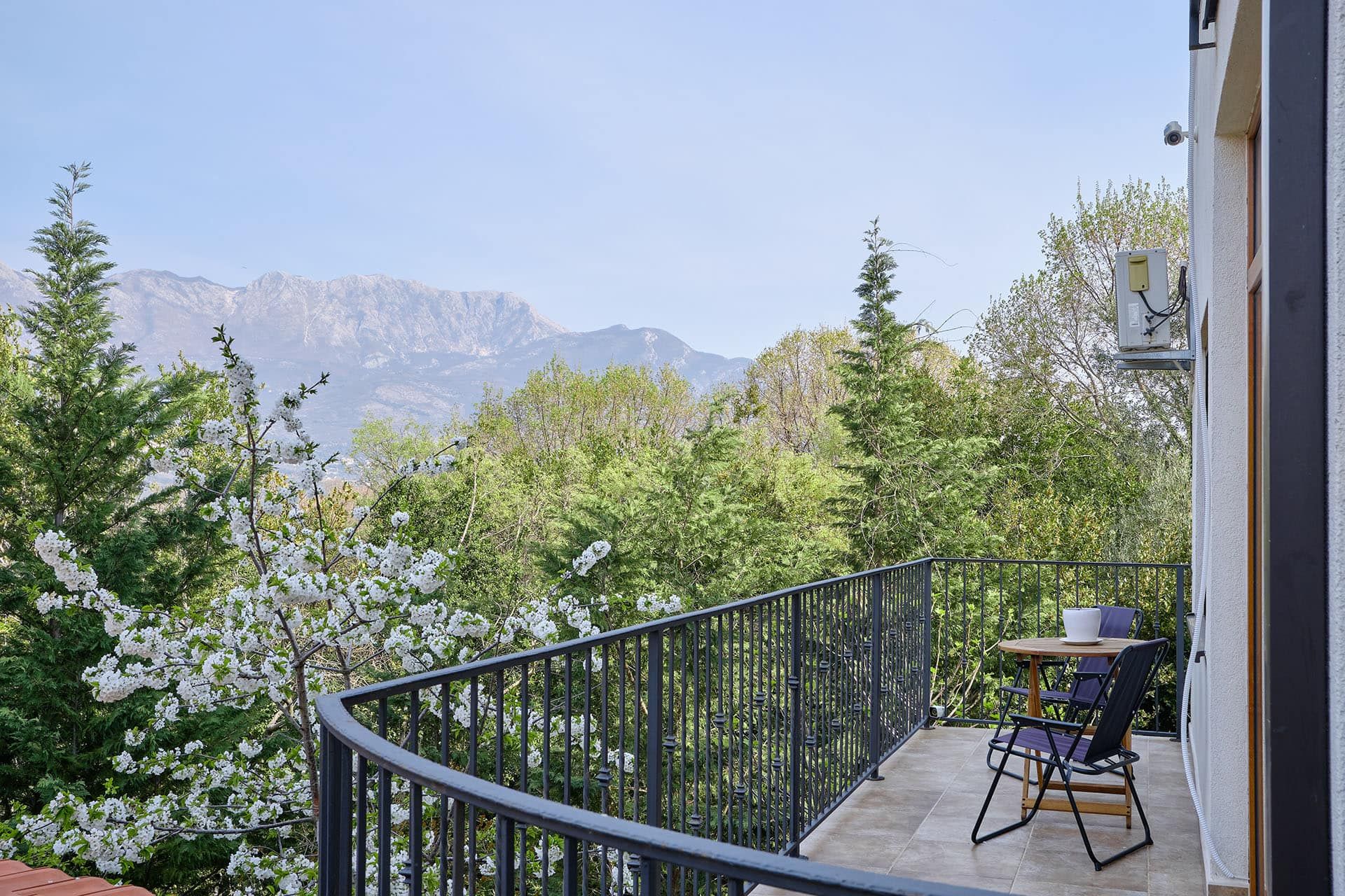 Balcony with black metal railing, small table and chairs overlooking mountains and blooming trees.
