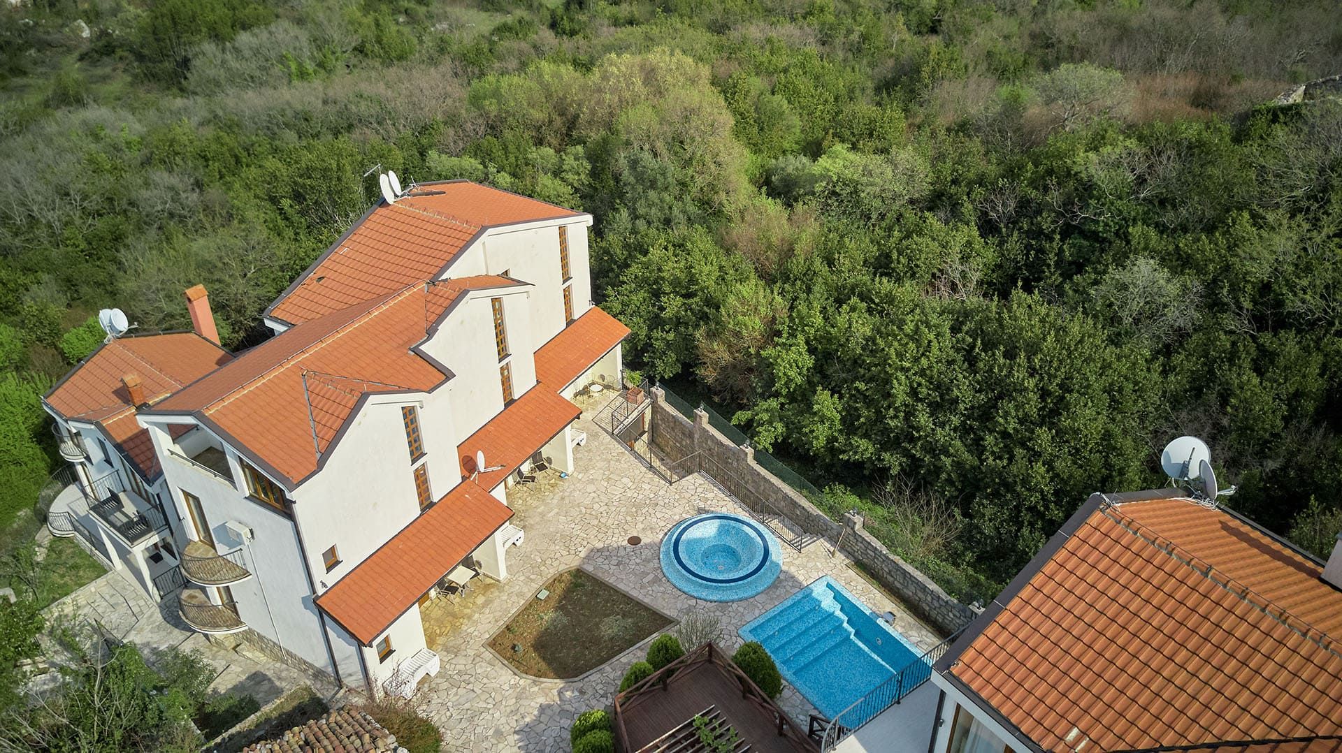 Aerial view of a house with a terracotta roof, stone courtyard, pool and jacuzzi surrounded by dense forest.