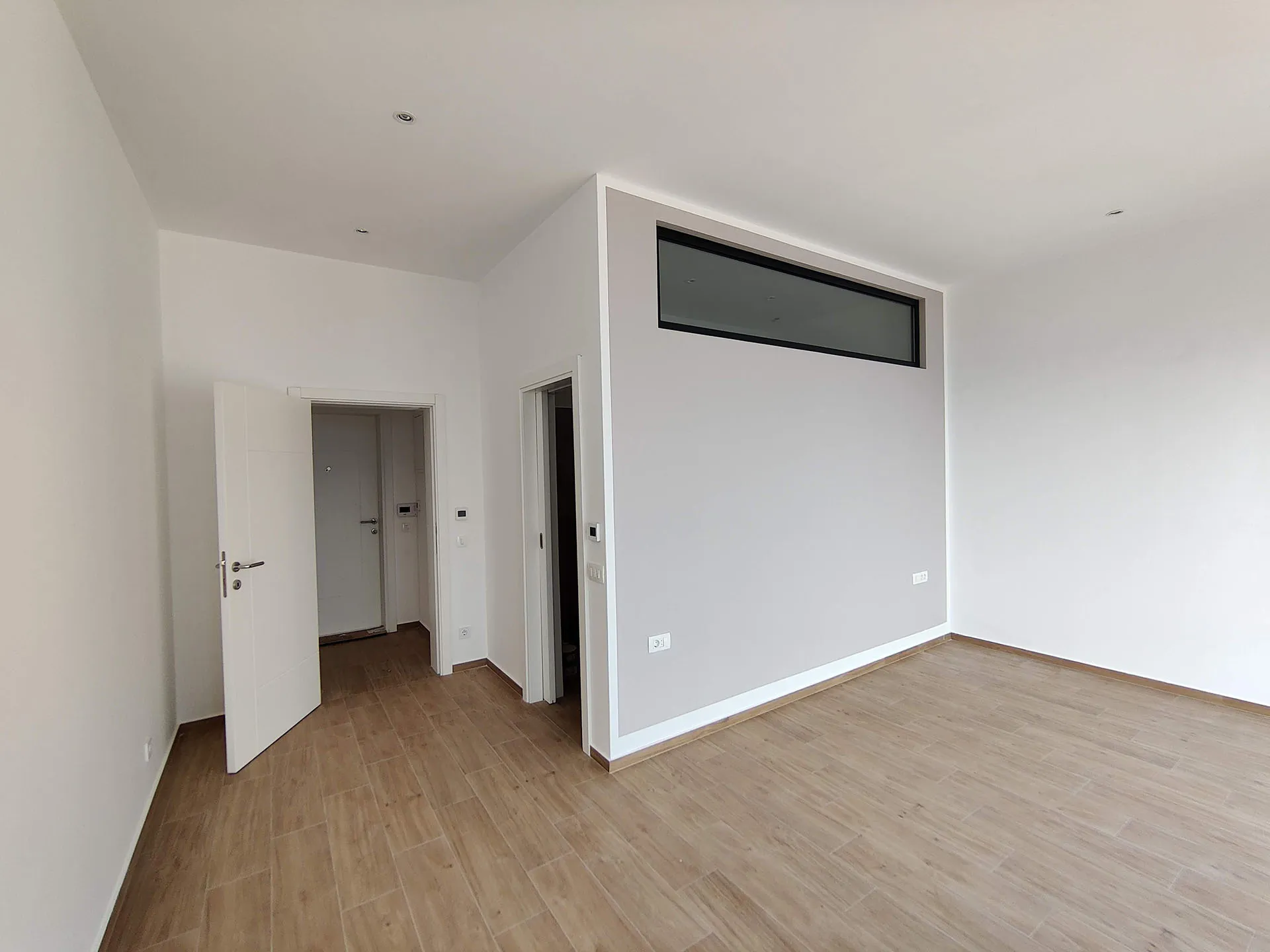 Empty room with laminate flooring, white walls, an open entry door, and a high horizontal window.