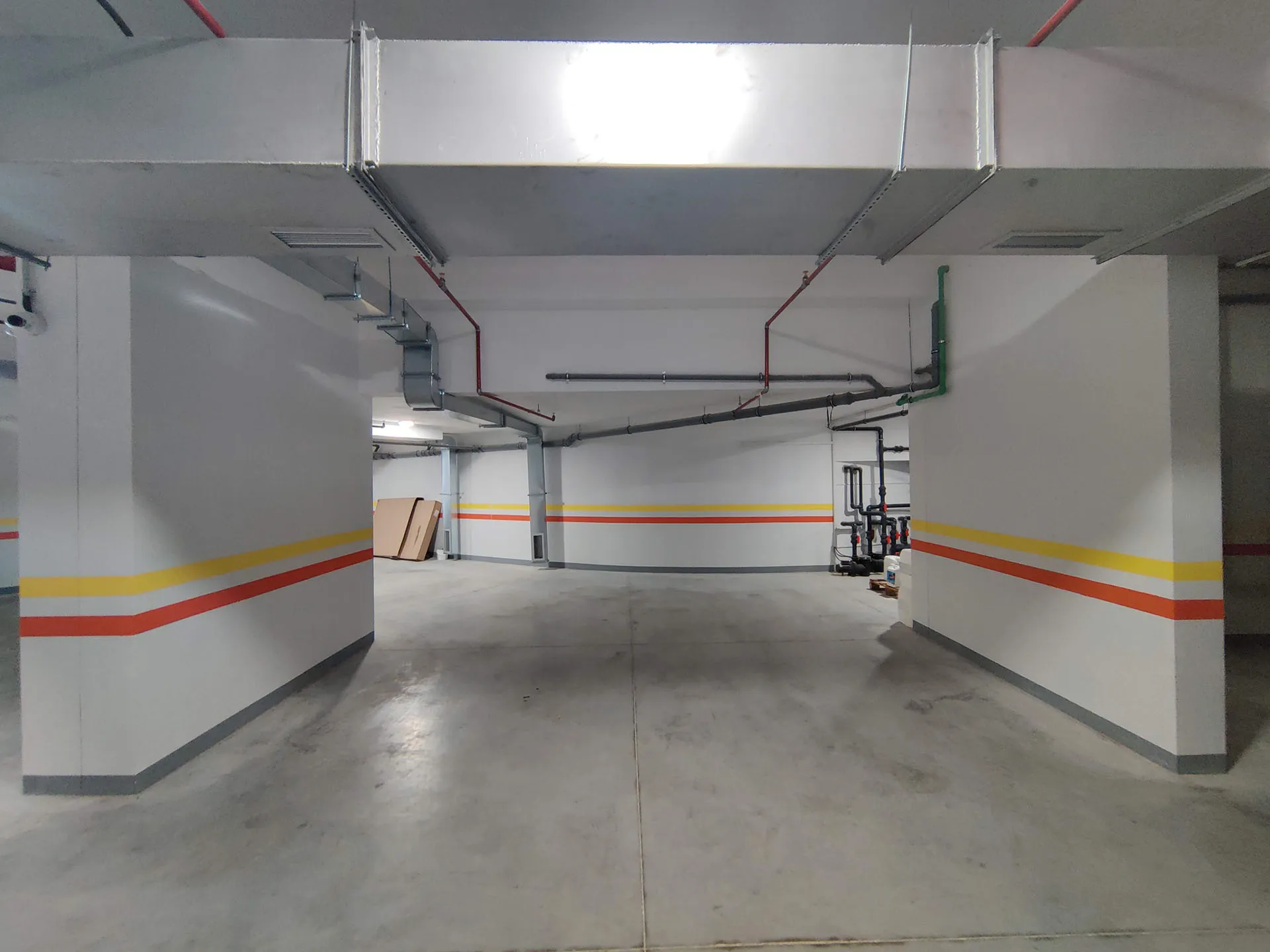 Underground parking or utility space with concrete floors, white walls featuring colored stripes, and exposed pipes on the ceiling.