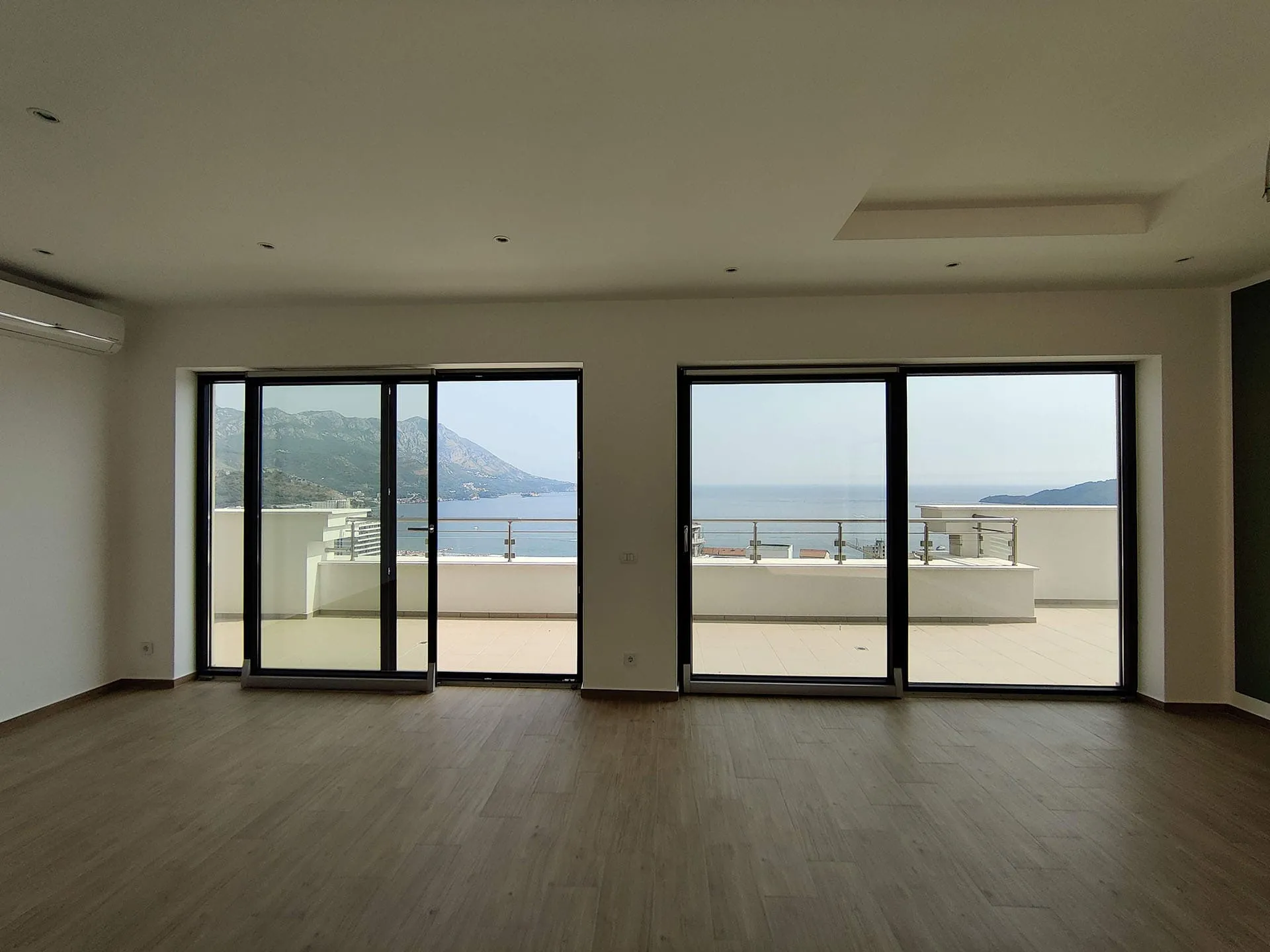 Empty room with wood flooring and large sliding glass doors opening to a balcony overlooking the sea and mountains.