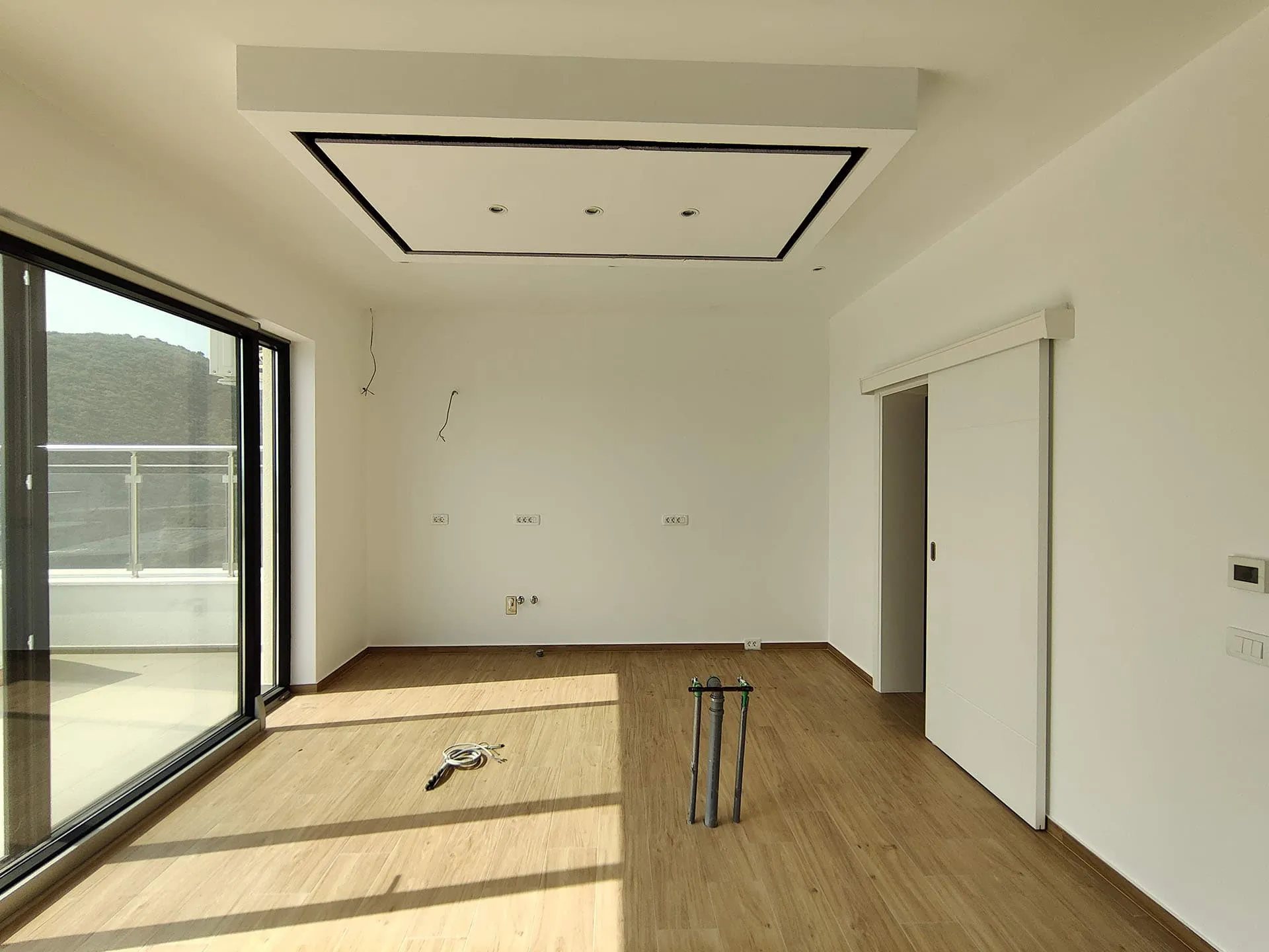 Empty room with wood flooring, large sliding glass door to balcony, and construction pipes on the floor.