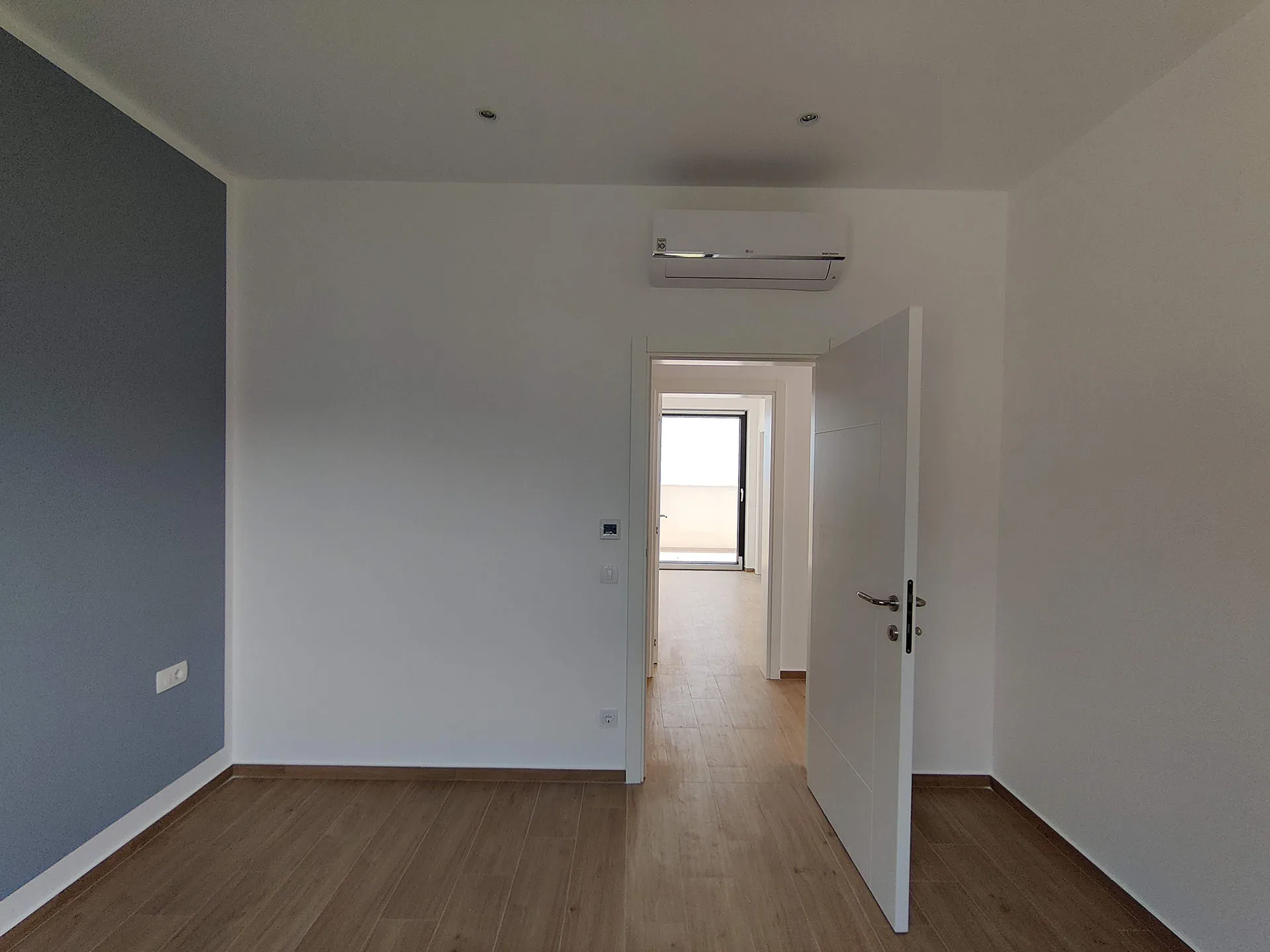 Empty room with wood flooring, a grey accent wall, and an air conditioner. An open door leads to a hallway.