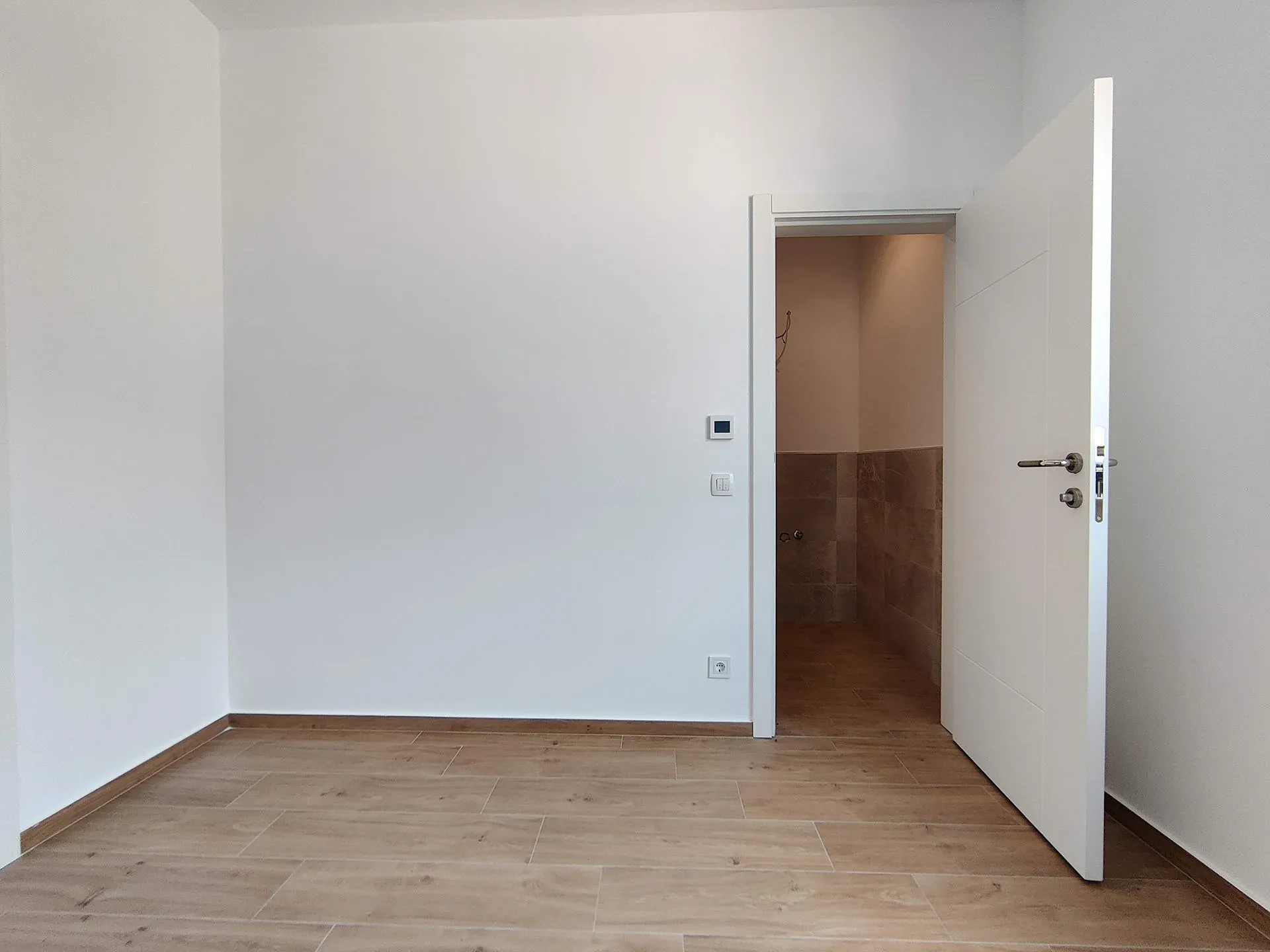 Empty room with white walls and light wood-look flooring. An open door leads to a tiled hallway or bathroom area.