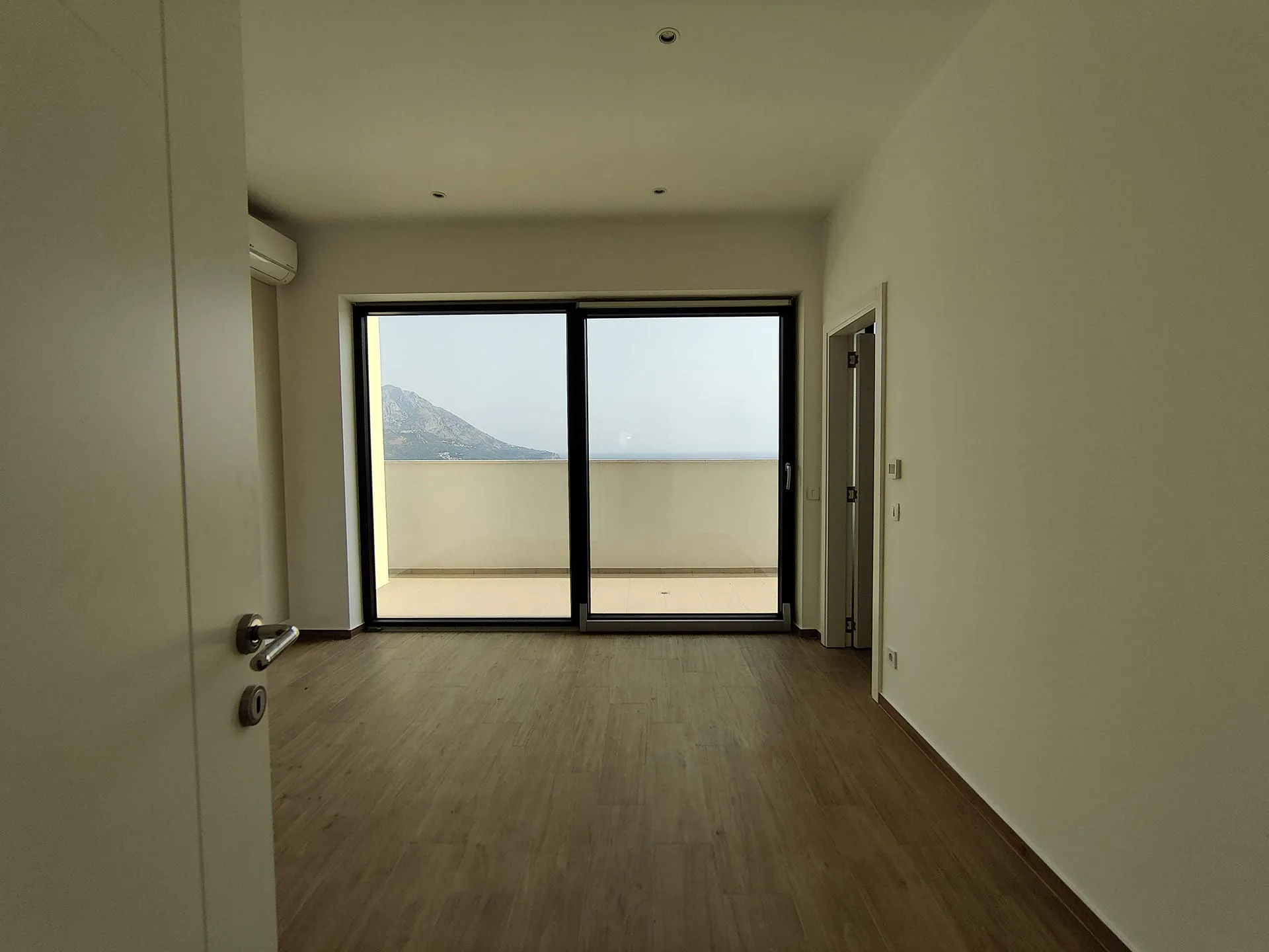 Empty room with wood flooring and large sliding glass doors opening to a balcony overlooking the sea and mountain.