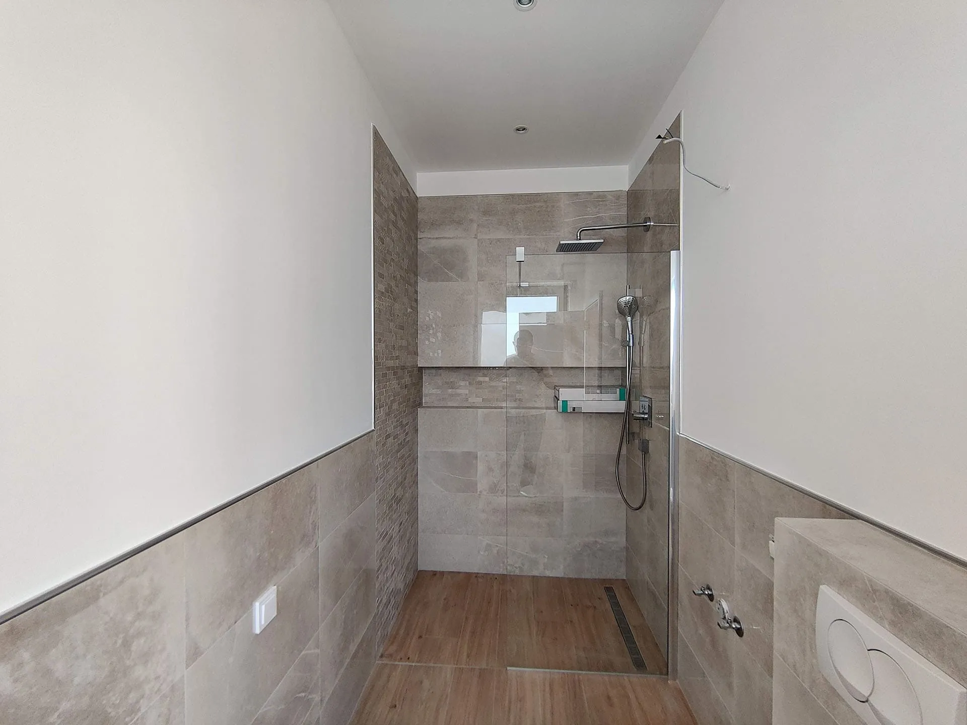 Modern bathroom featuring a walk-in shower with glass partition, grey tiles, wood-look flooring, and chrome fixtures.