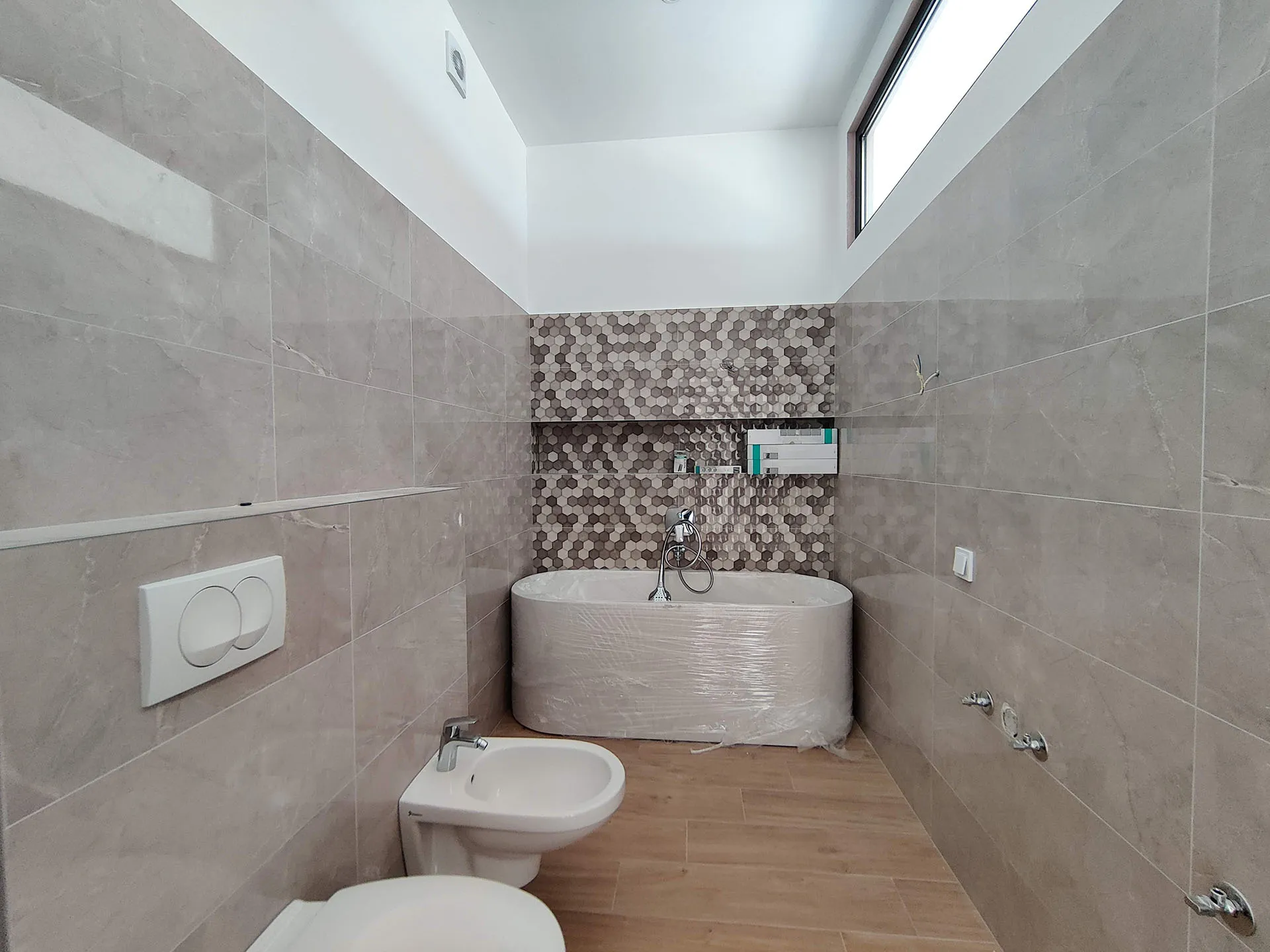 Modern bathroom with gray tiles, a plastic-wrapped freestanding bathtub, bidet, and toilet.