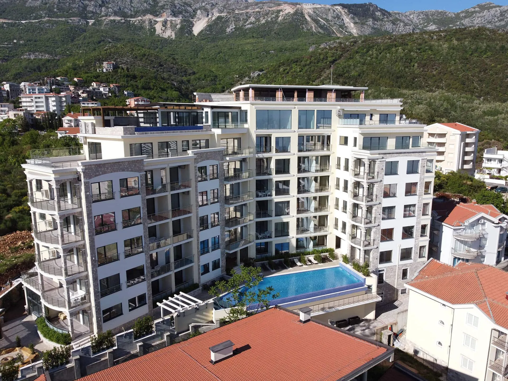 Aerial view of a multi-story apartment complex with a pool and balconies against mountains.