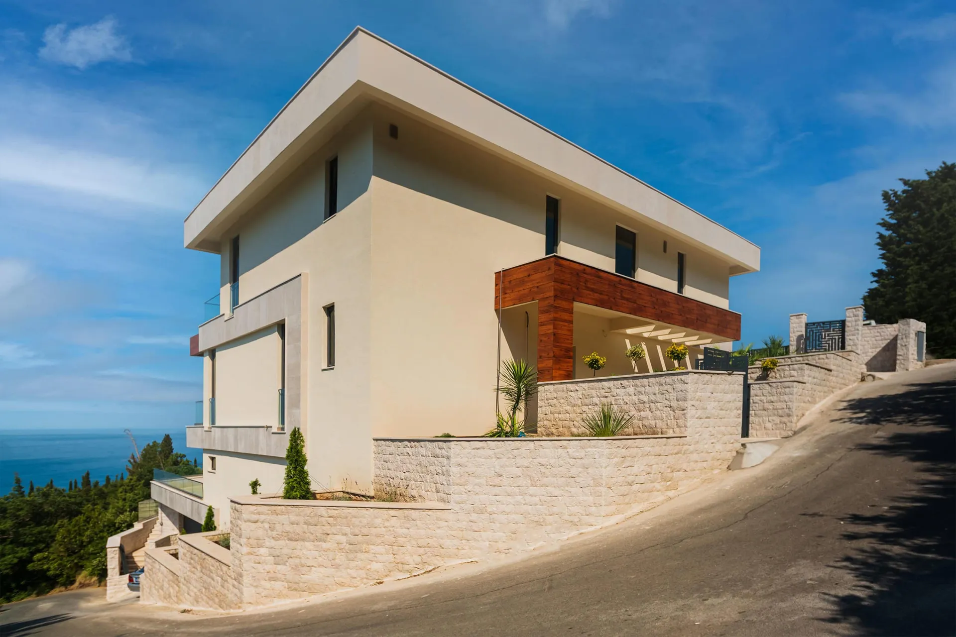 Modern two-story house with beige walls and wooden balcony cladding on a hillside overlooking the sea.