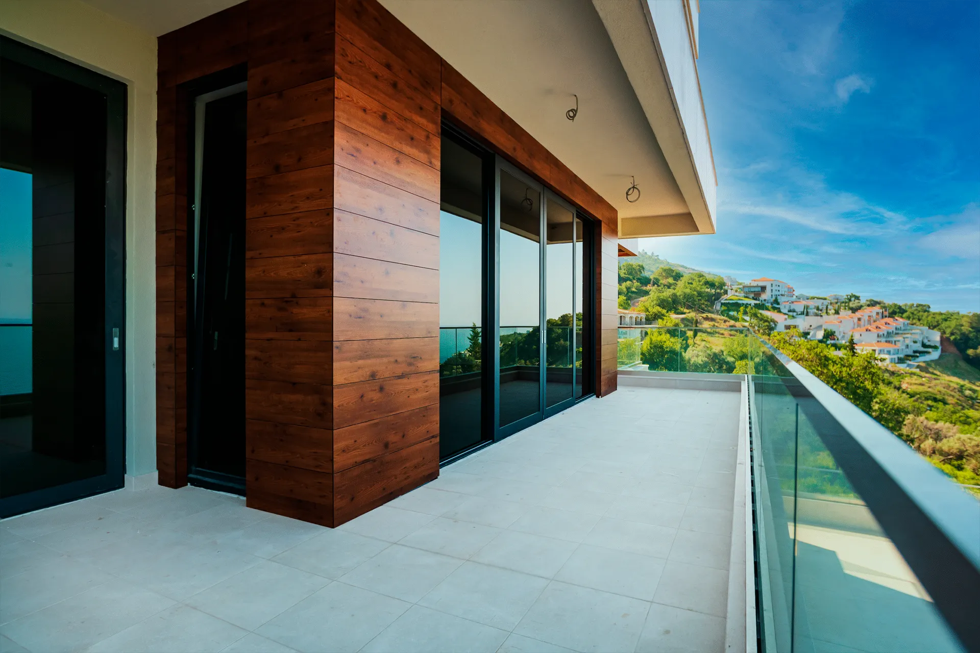 Wide balcony with wood wall and glass railing overlooking hills and white houses.