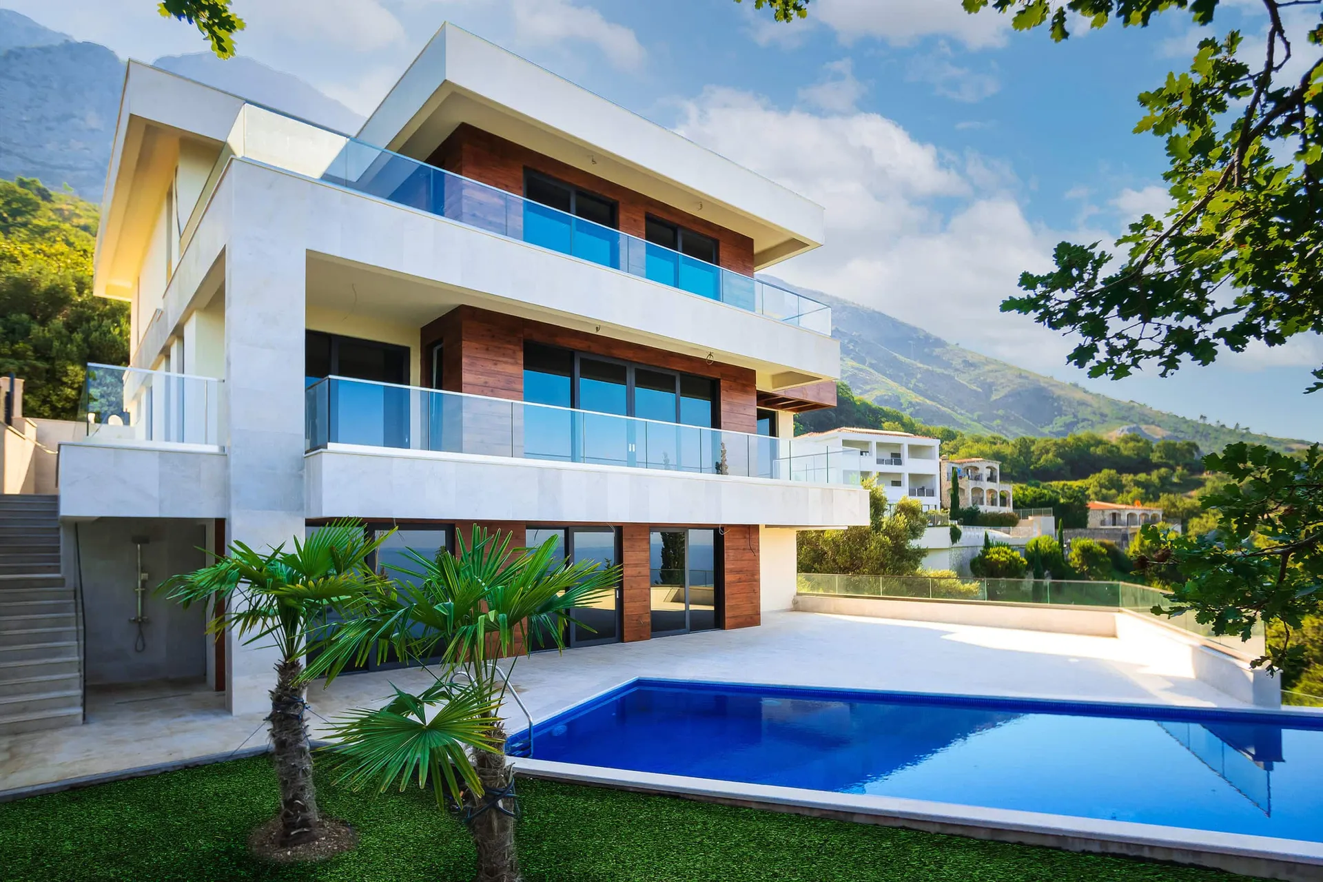 Modern multi-story house exterior with a blue pool, glass balconies, and palm trees against mountains.