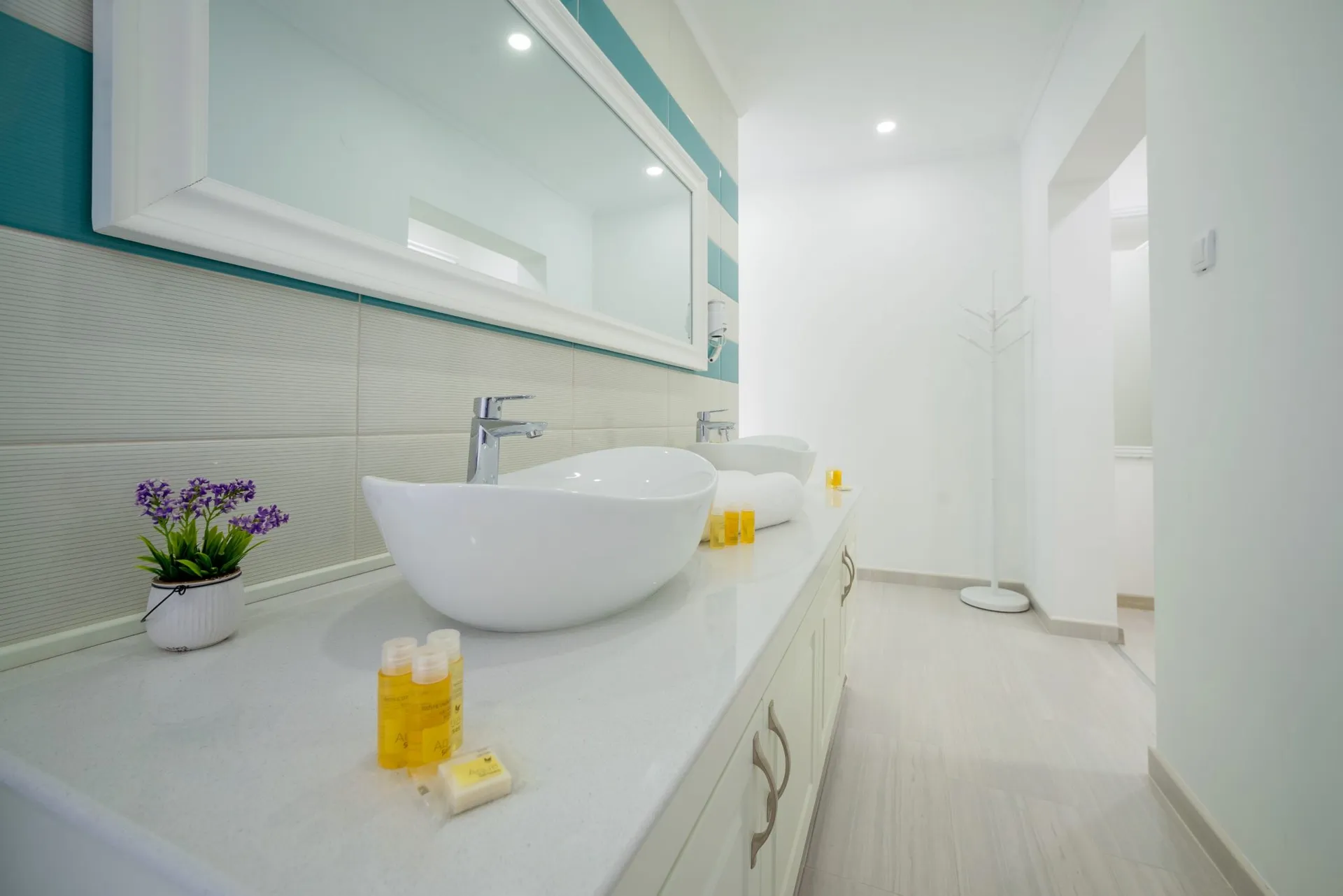 Modern bathroom featuring two white vessel sinks, a large framed mirror, toiletries, and teal accent wall tiles.