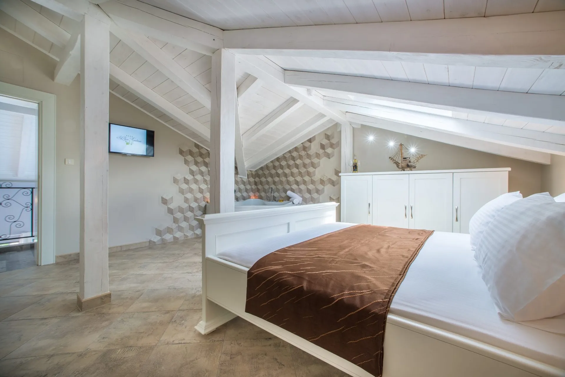 Bright attic bedroom with a white wooden bed, exposed beams, and an open bathtub visible in the background.