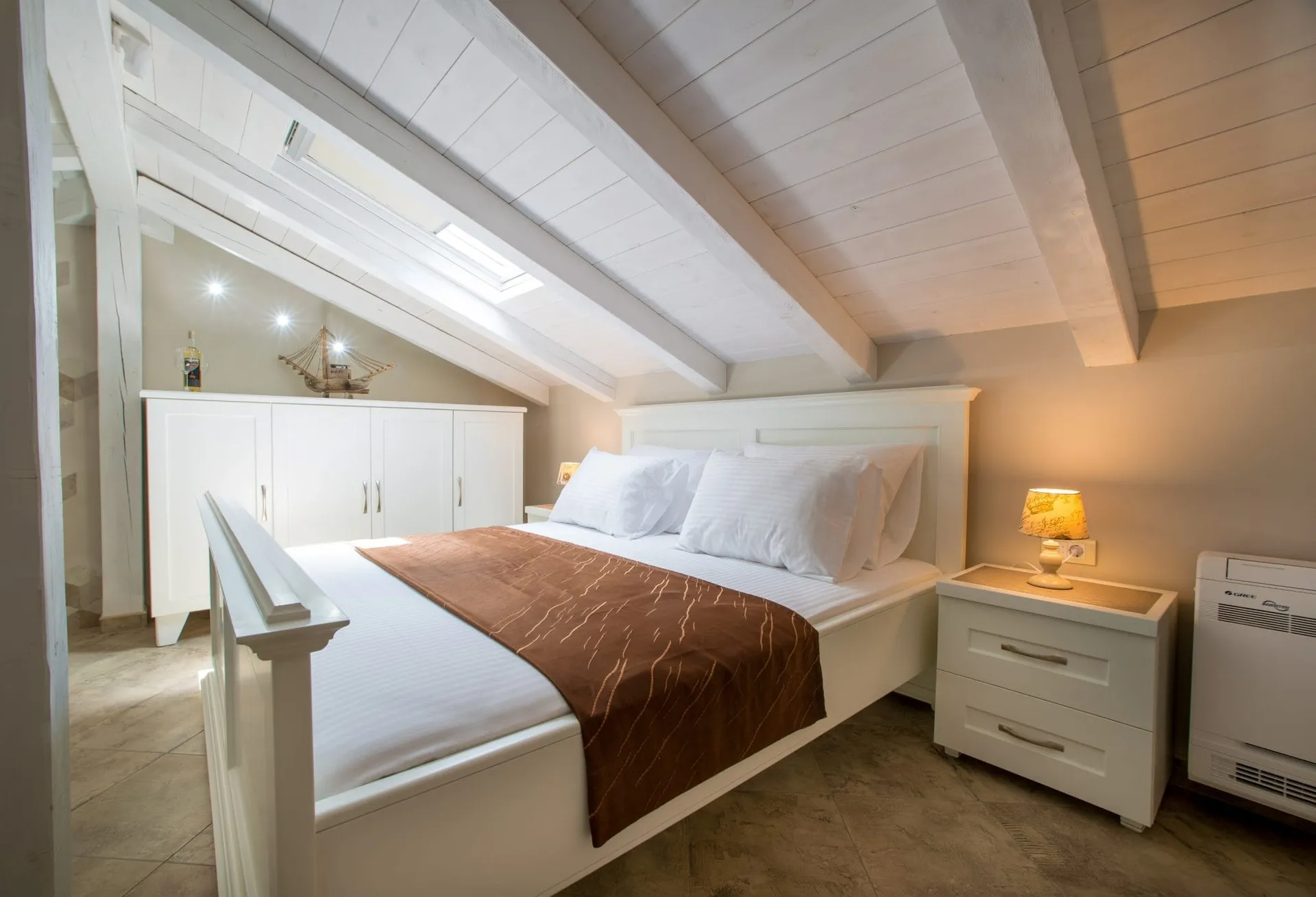 Bright attic bedroom featuring a white wooden bed, wardrobe, sloped ceiling with beams and skylight.